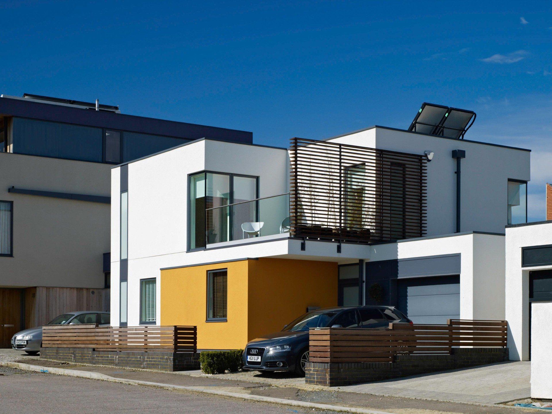 Modern houses with white and yellow facades, wooden fences, and parked cars under a clear blue sky