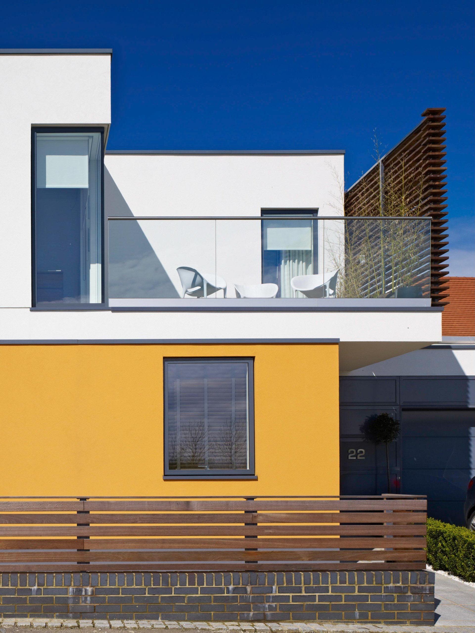 Modern two-story house with white and mustard walls, blue sky, balcony, and wooden fence