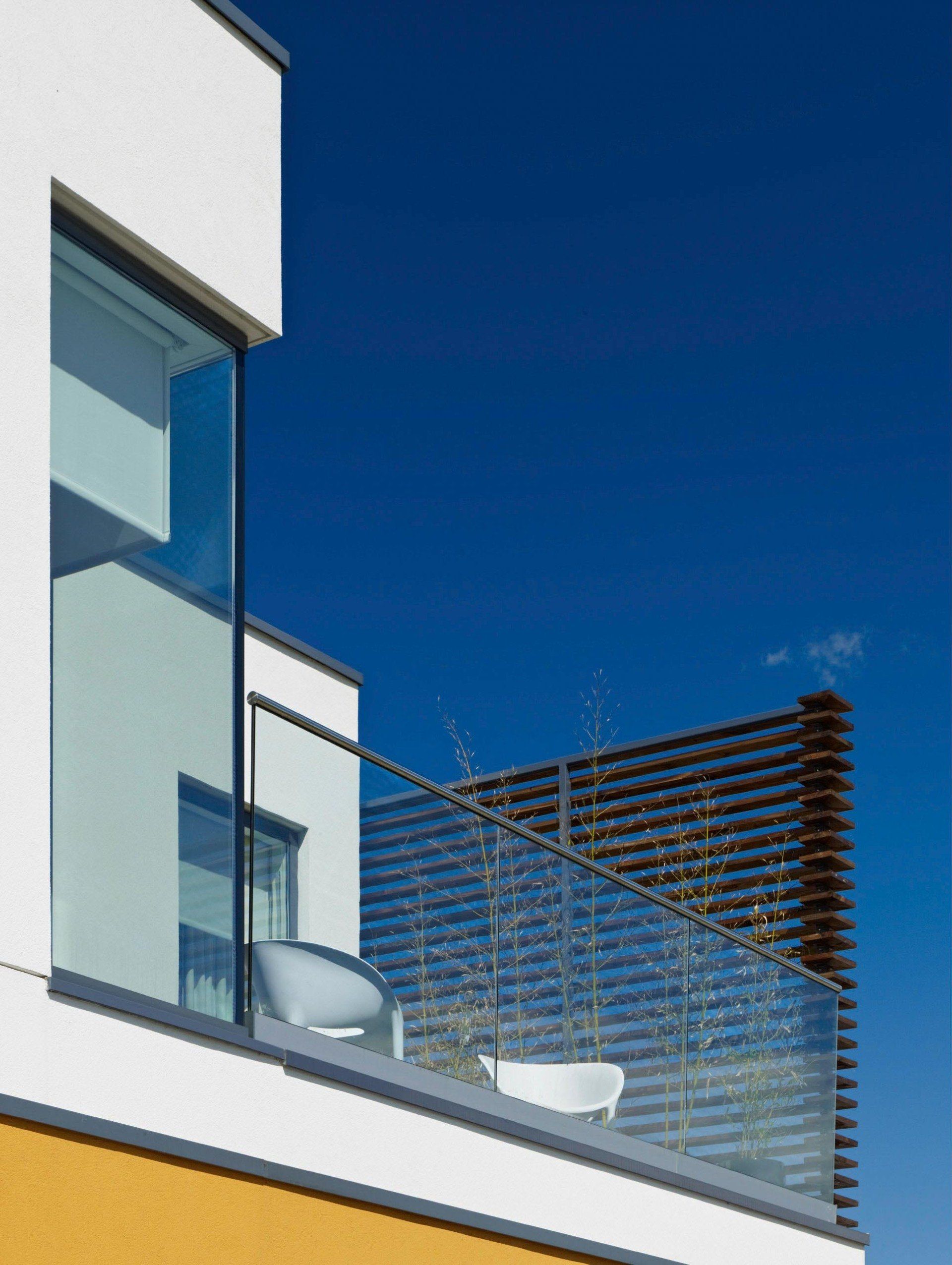 Modern apartment balcony with glass railing and white chairs against a clear blue sky