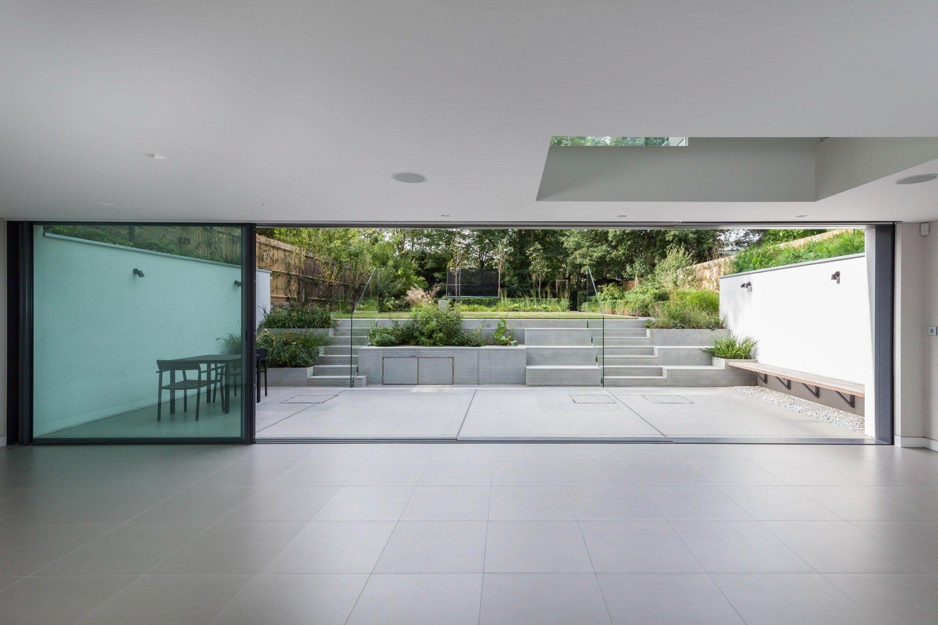 Modern open-plan patio with gray tiles, glass walls, and steps leading to a landscaped garden