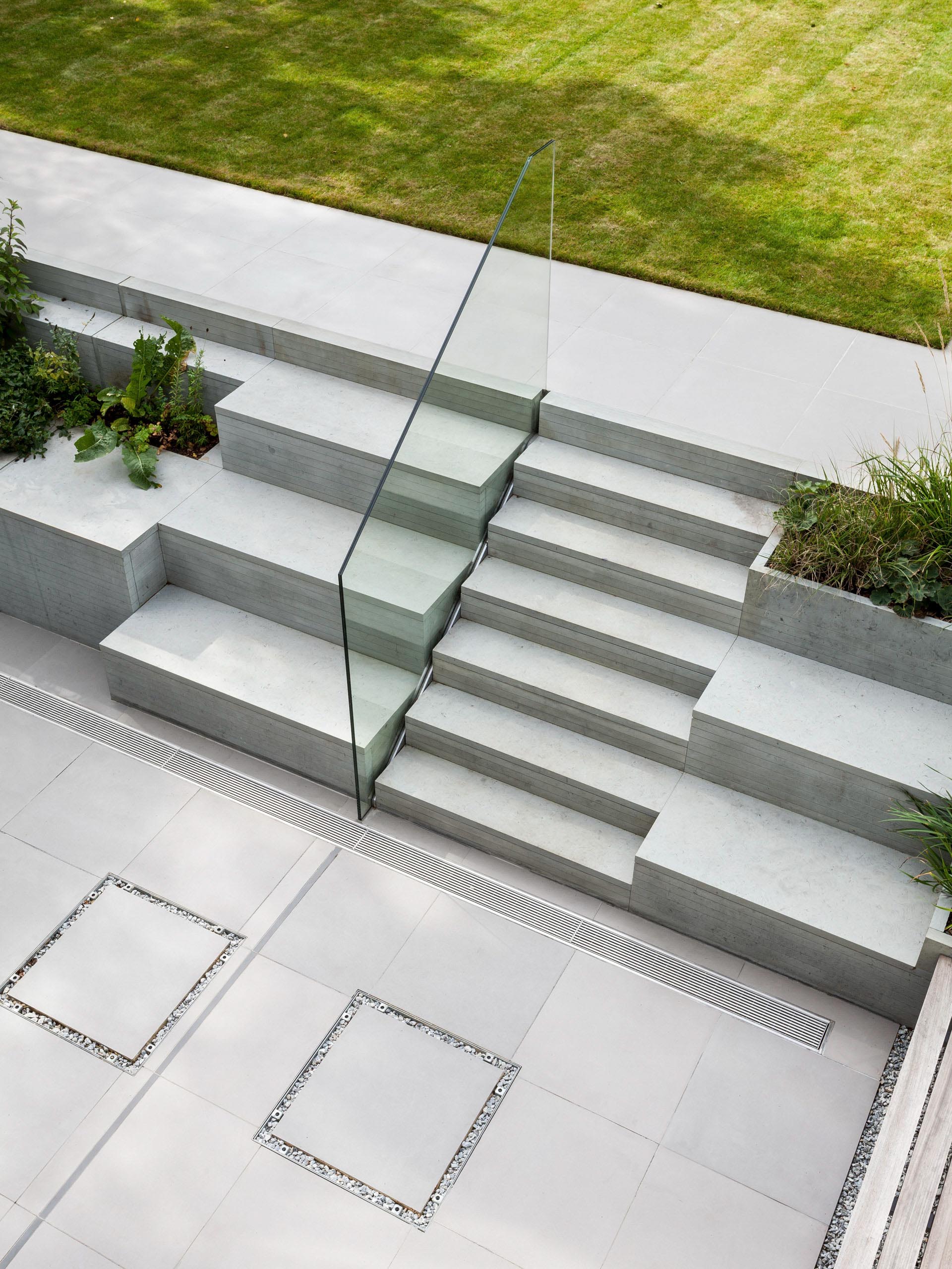 Modern outdoor staircase with glass railing beside a green lawn and paved patio