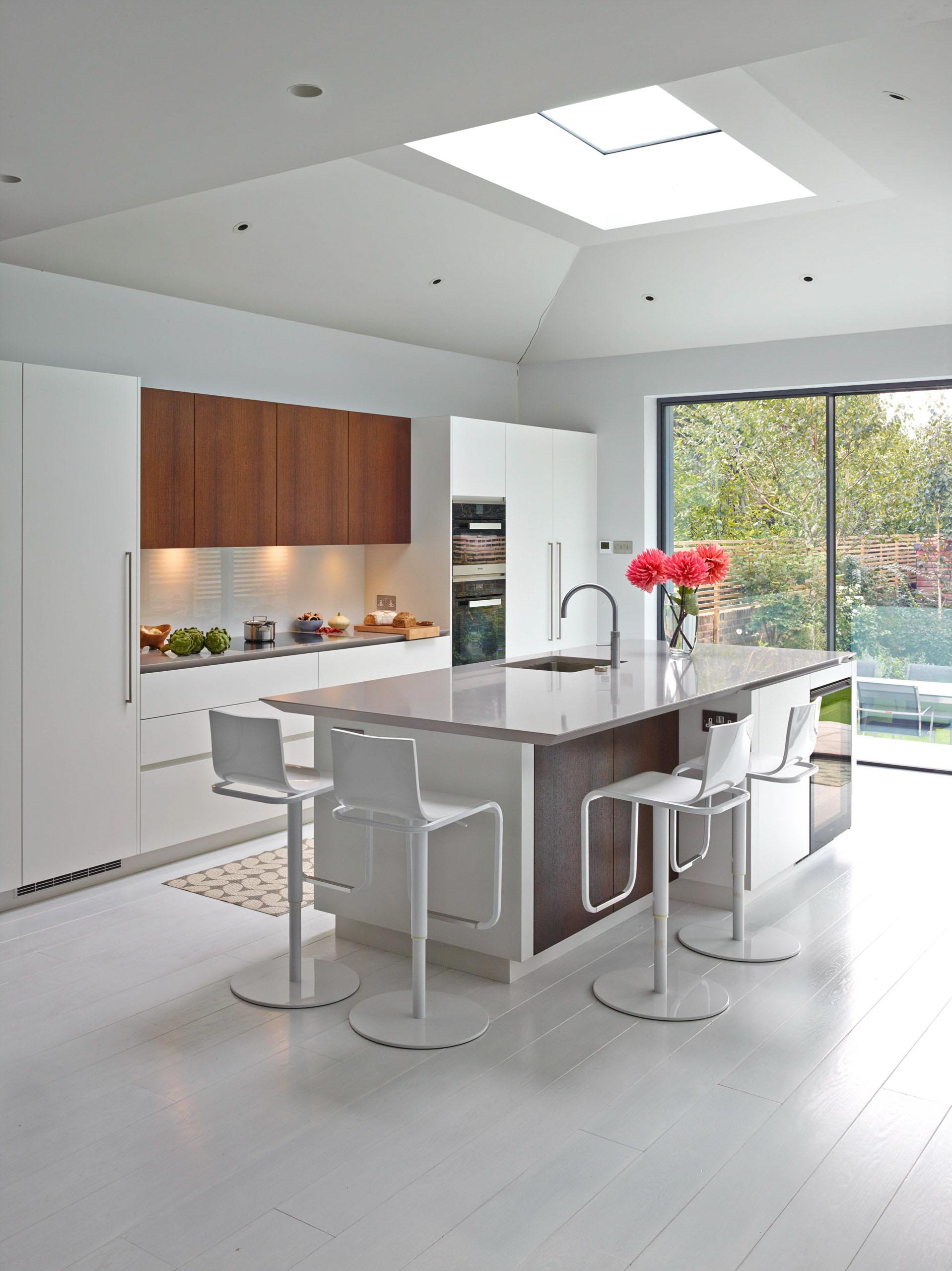 Modern white kitchen with island, wood accents, and skylight. Sliding glass doors lead to a garden.