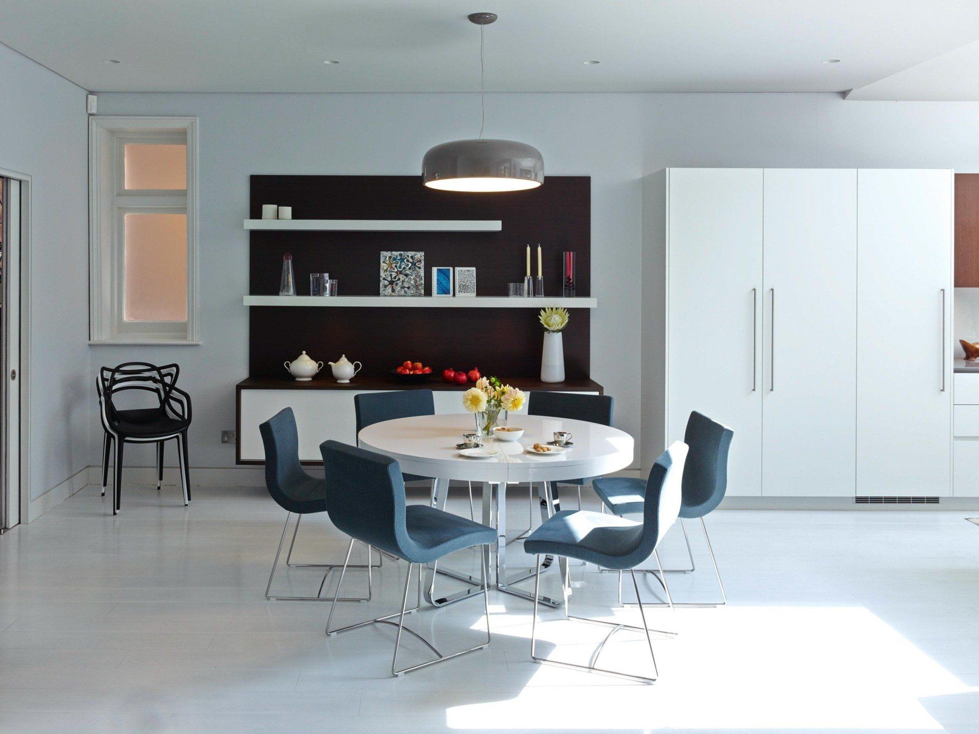 Modern dining room with round white table, blue chairs, dark shelving, and large white cabinet