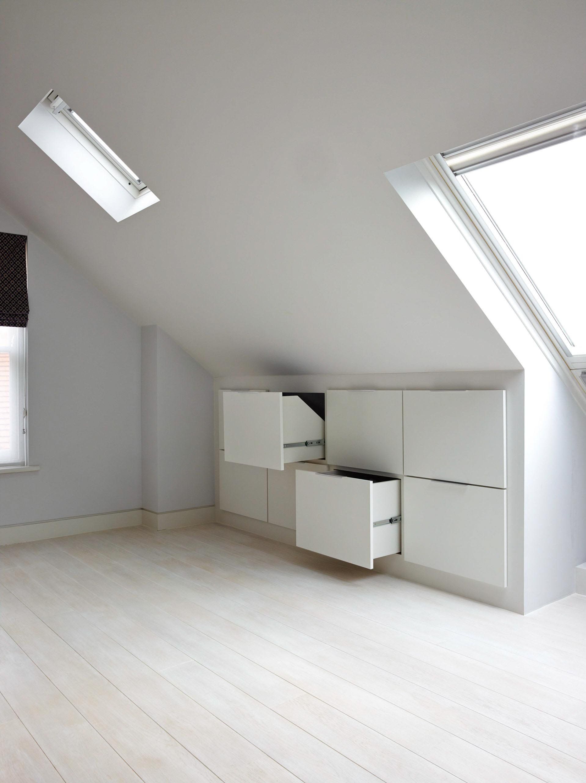 Bright, empty attic room with white walls, slanted ceiling, and built-in white cabinets. Sunlight streams in from windows.