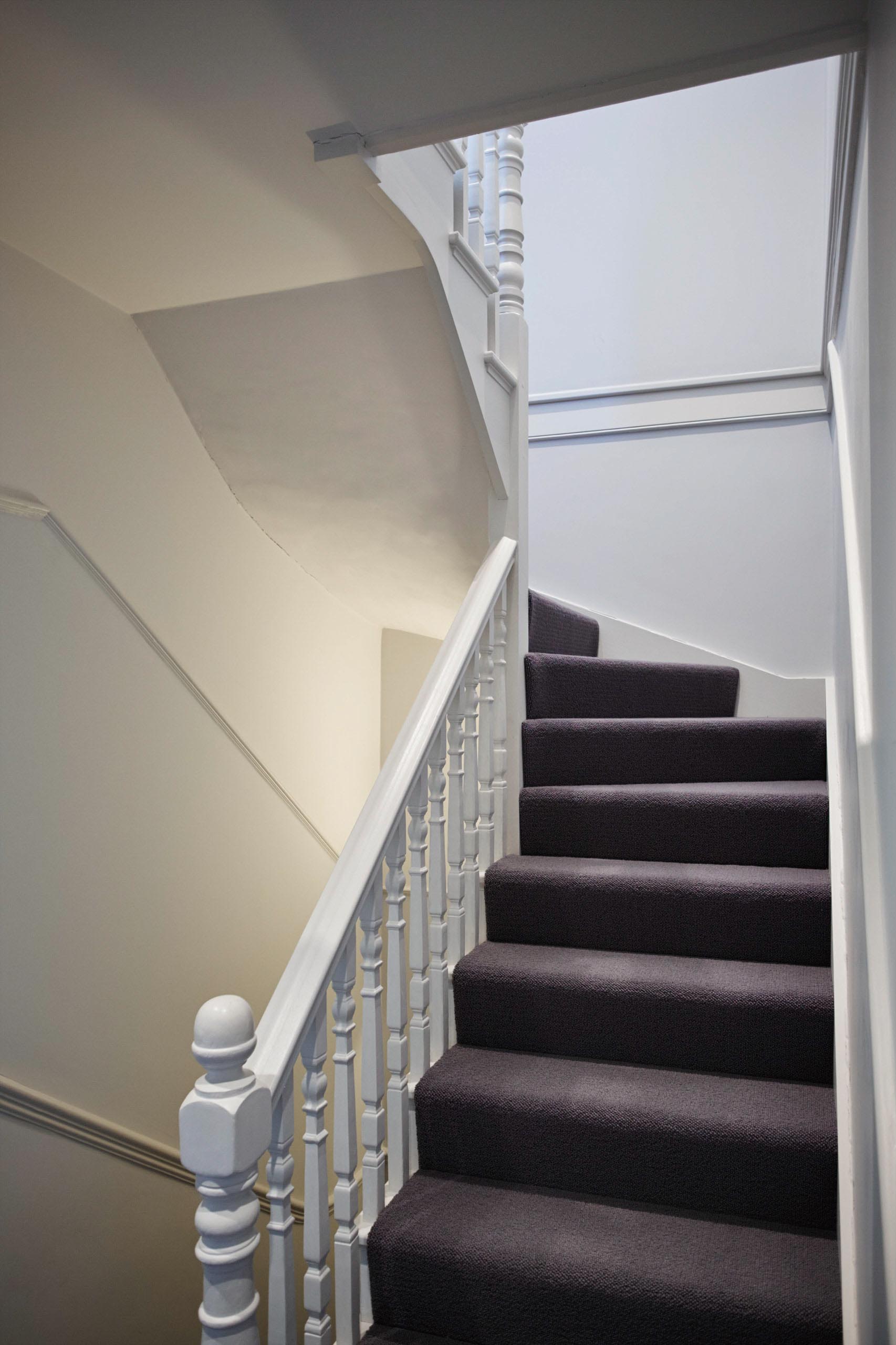 Staircase with dark carpet and white railing, leading upwards towards a bright opening.