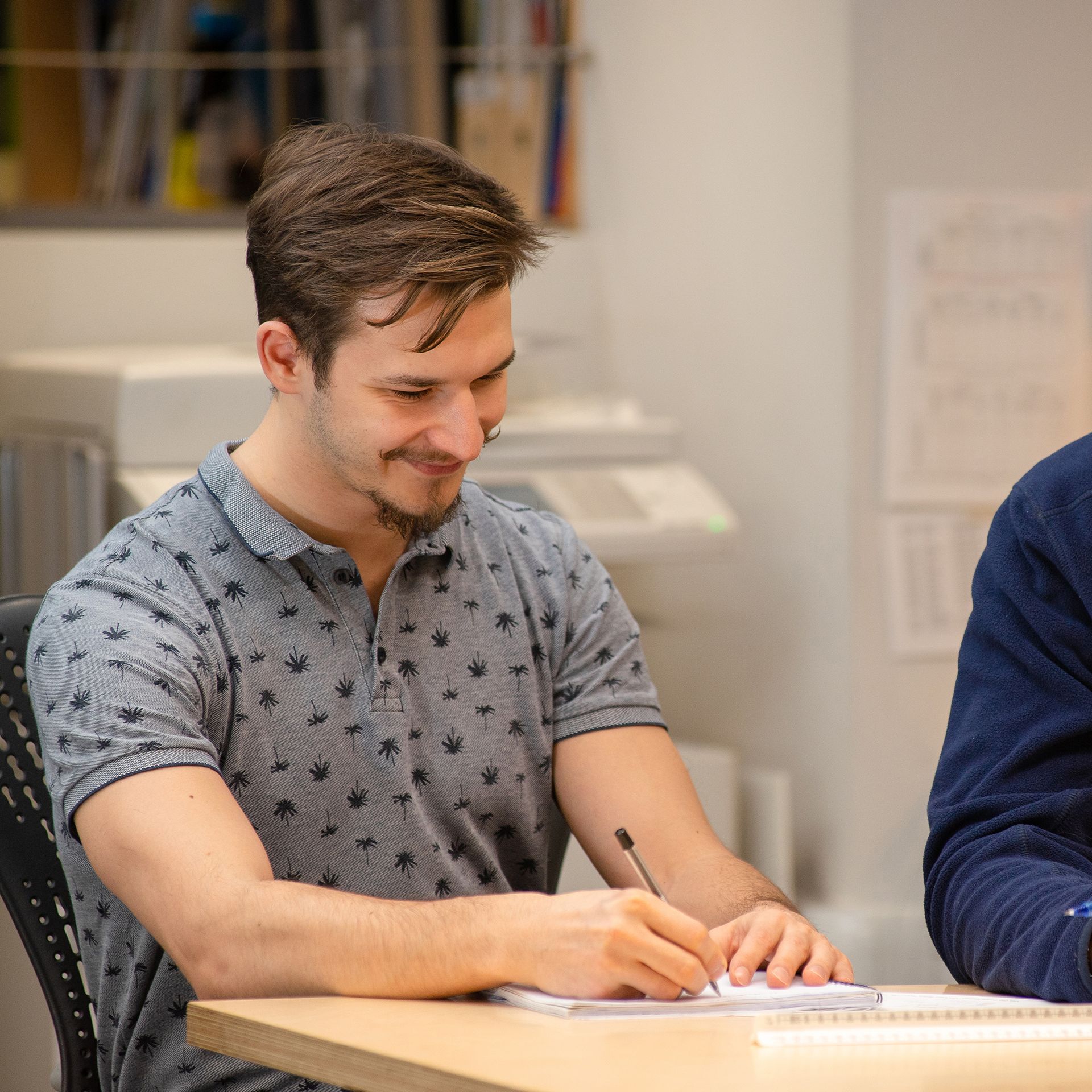 Man in a gray patterned shirt writing at a table in a meeting room