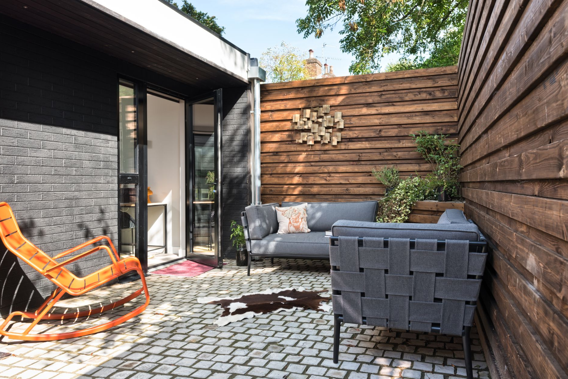 Modern patio with orange chair, gray sofa, planter, and wood-and-brick walls
