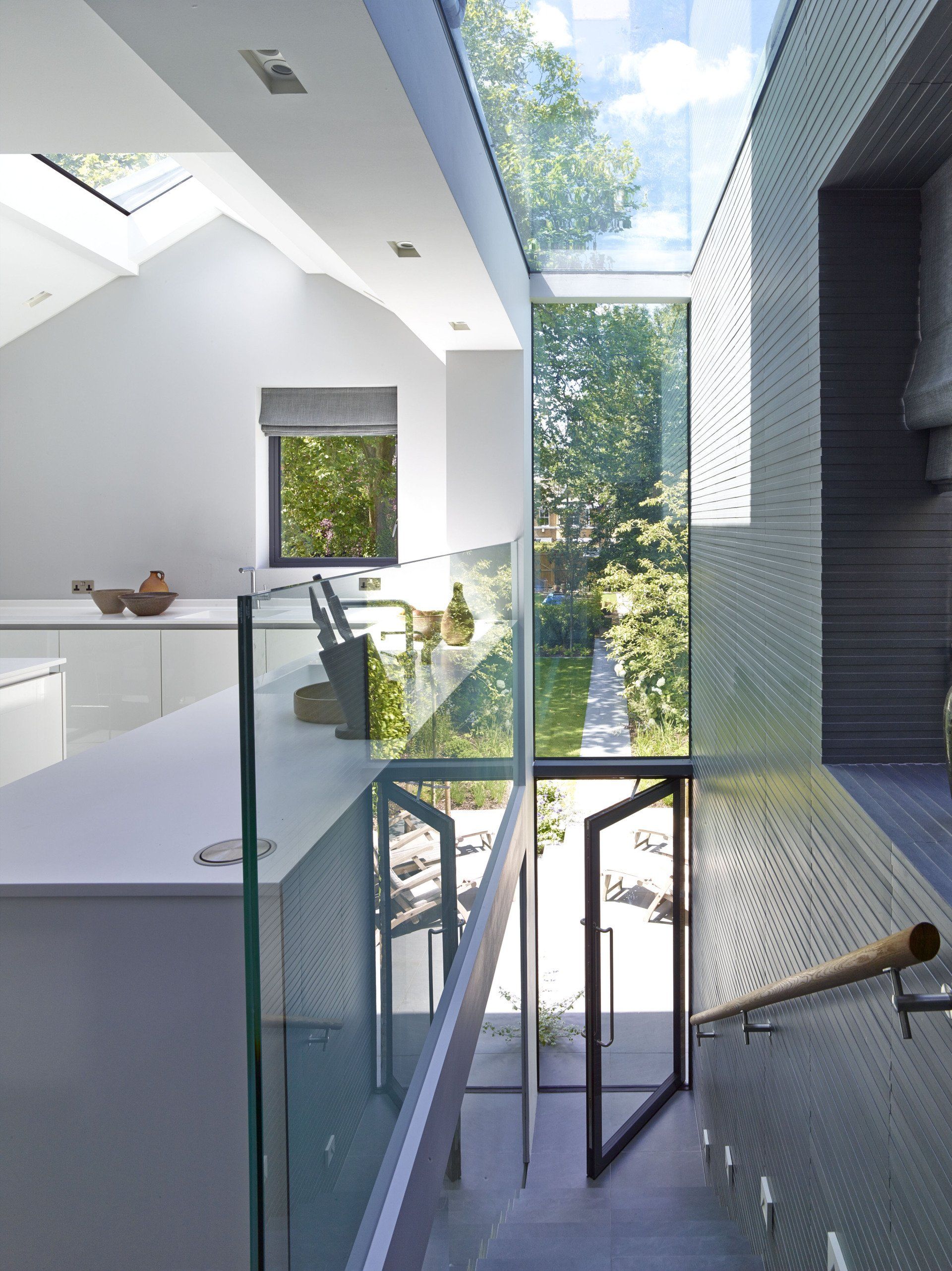 Modern white interior with stairs, glass railings, skylights, and greenery outside.