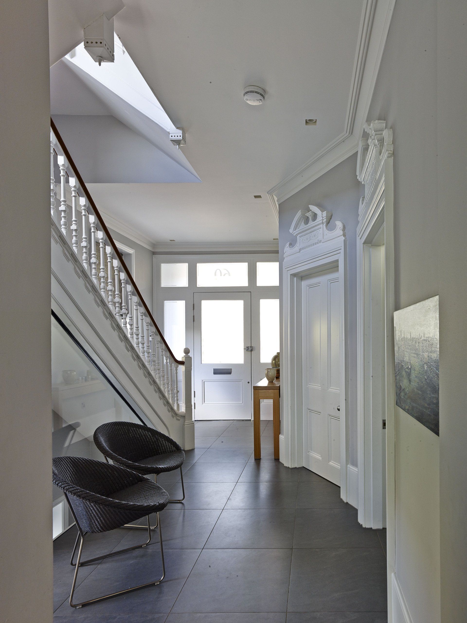 Bright white hallway with stairs, skylight, black chairs, and doors leading to a windowed room.