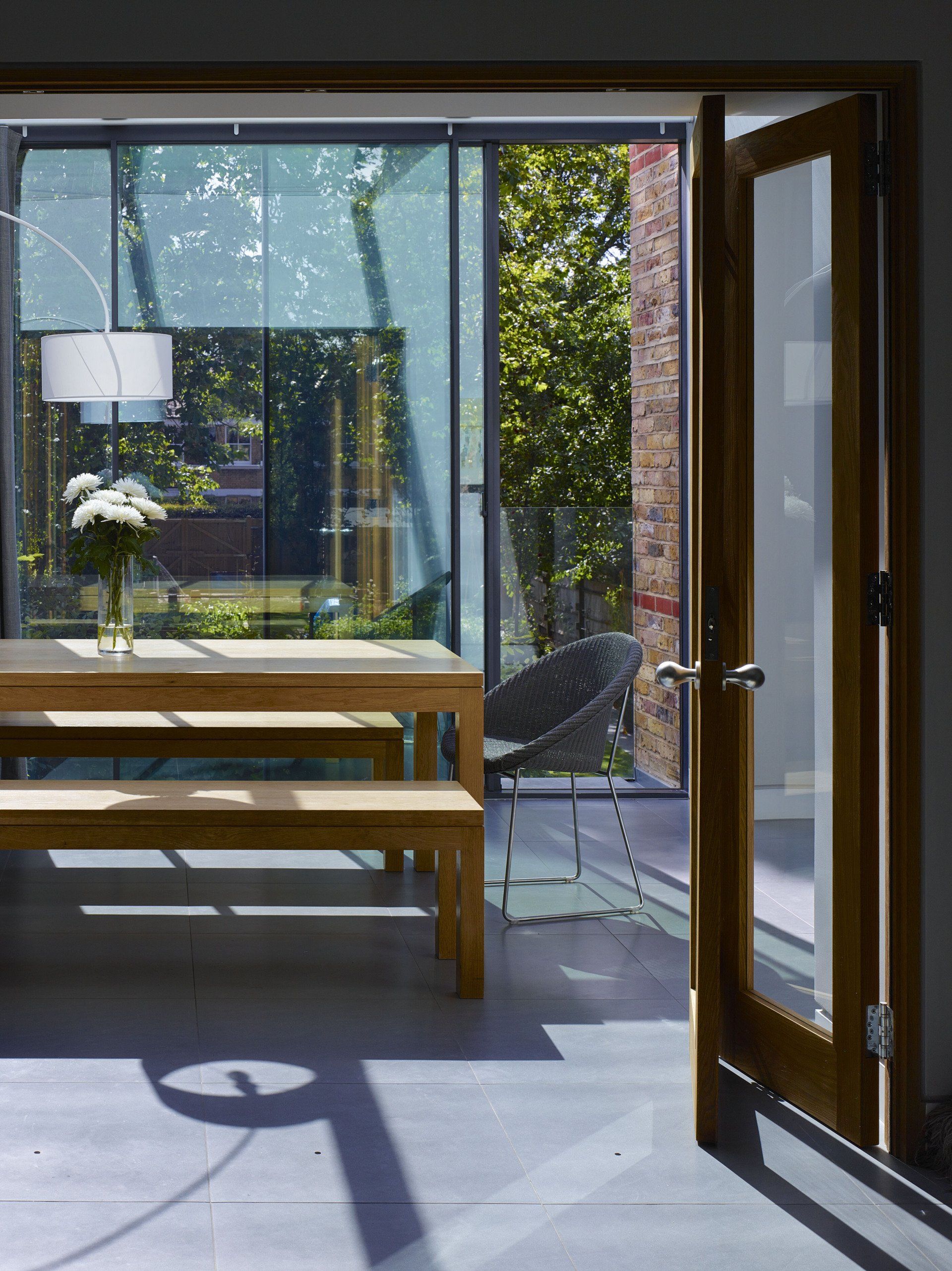 Bright room with wooden table by open glass doors, showing sunlight, plants, and an outdoor patio.