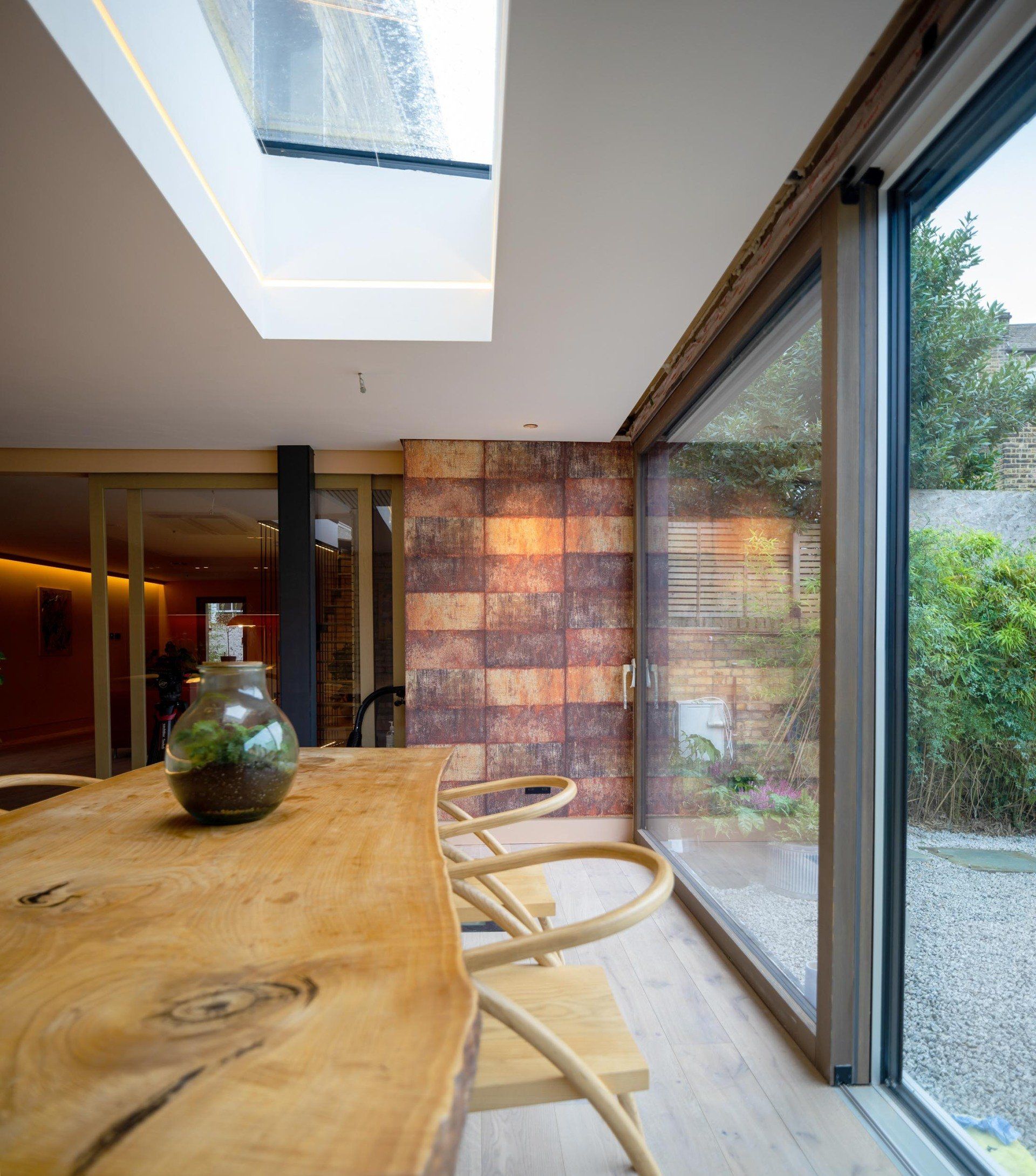Modern dining room with a live-edge wood table, chairs, skylight, and large sliding glass doors to a garden.