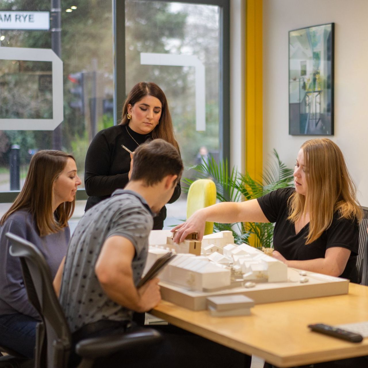 Four people discuss a tabletop model in a bright room with yellow trim and large windows.