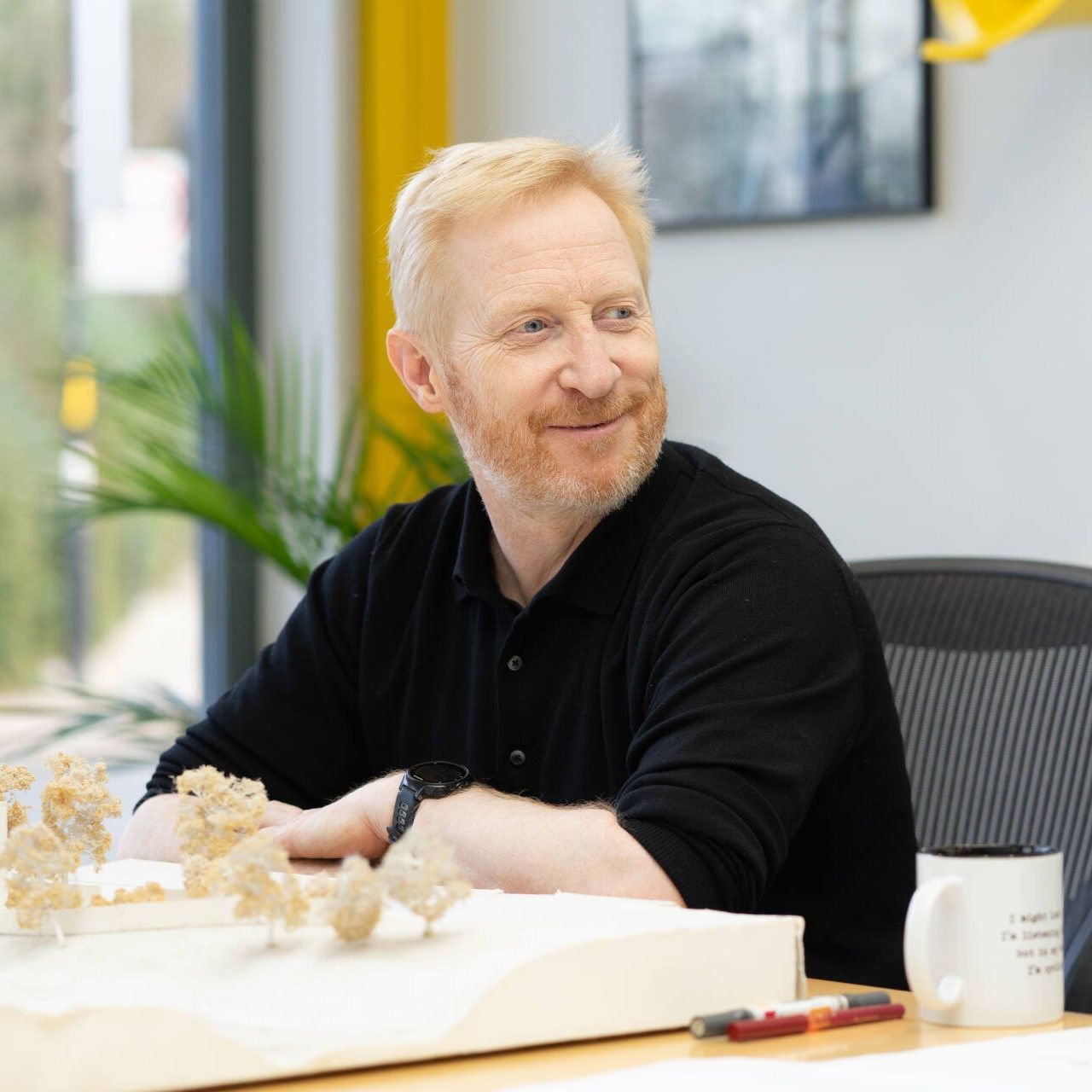 Man in black shirt smiling at a table with a model landscape and coffee mug in a bright office