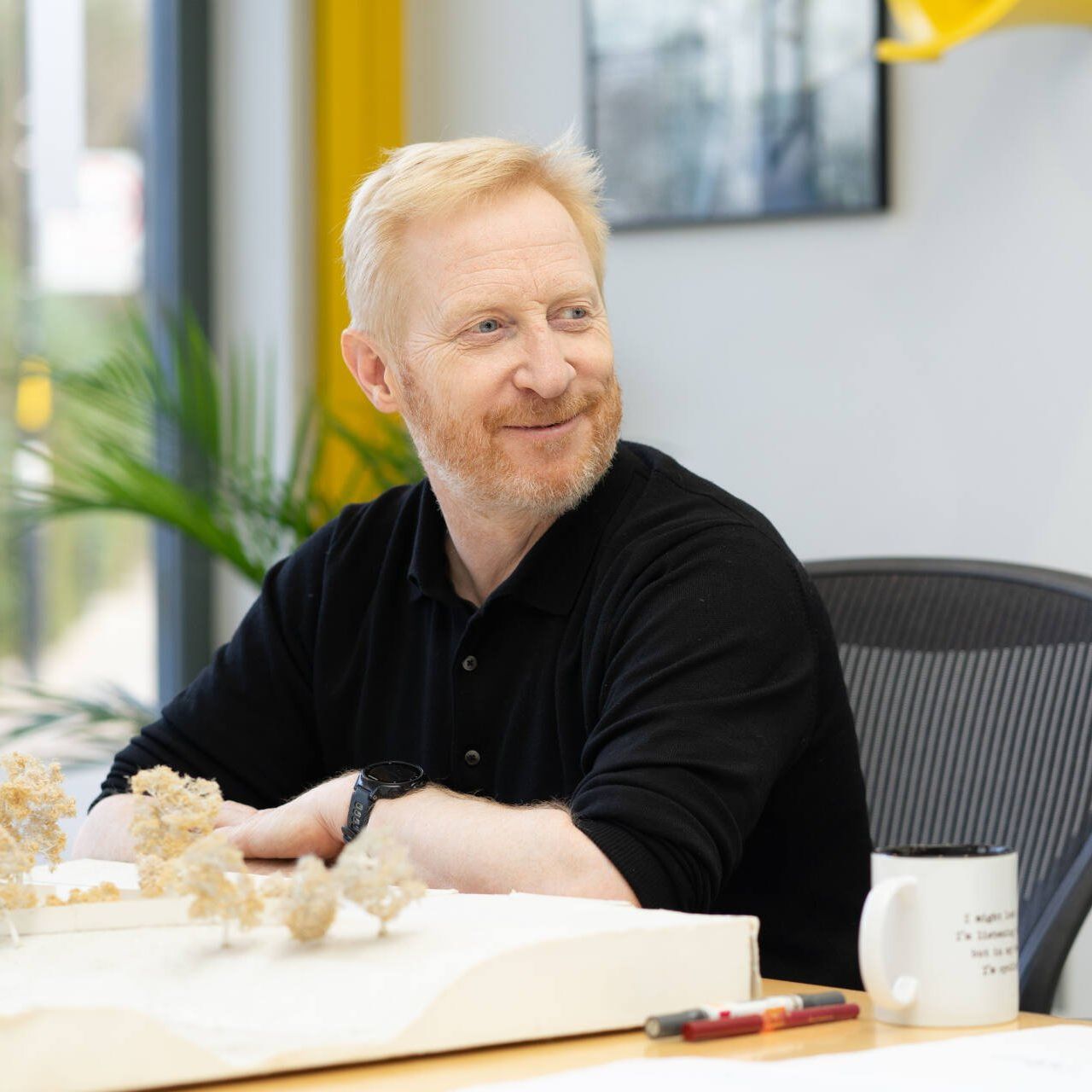 Smiling man in black shirt sitting at a desk with model pieces and a white mug in a bright office