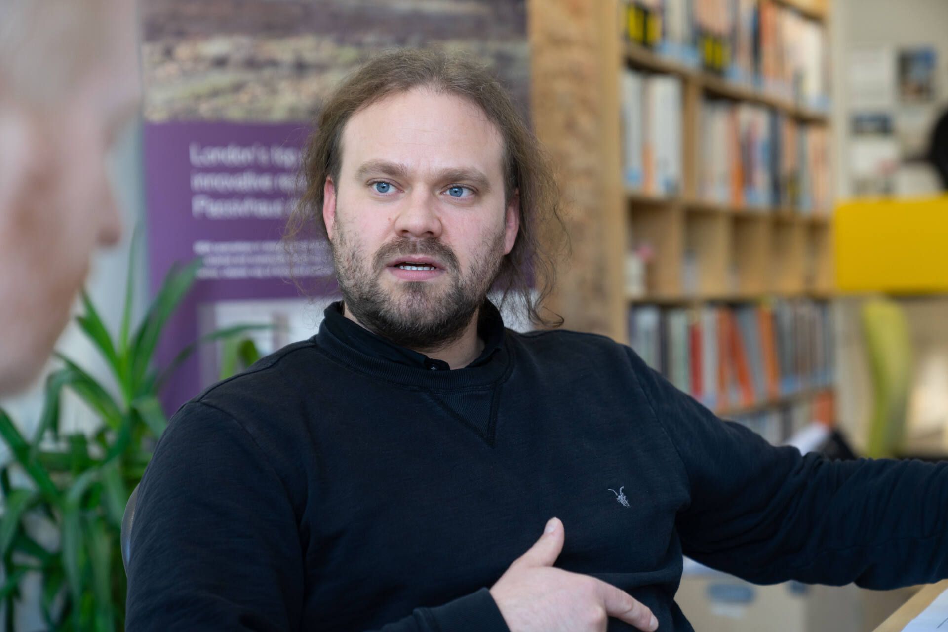 Man gesturing in a library, with bookshelves and a purple poster in the background.