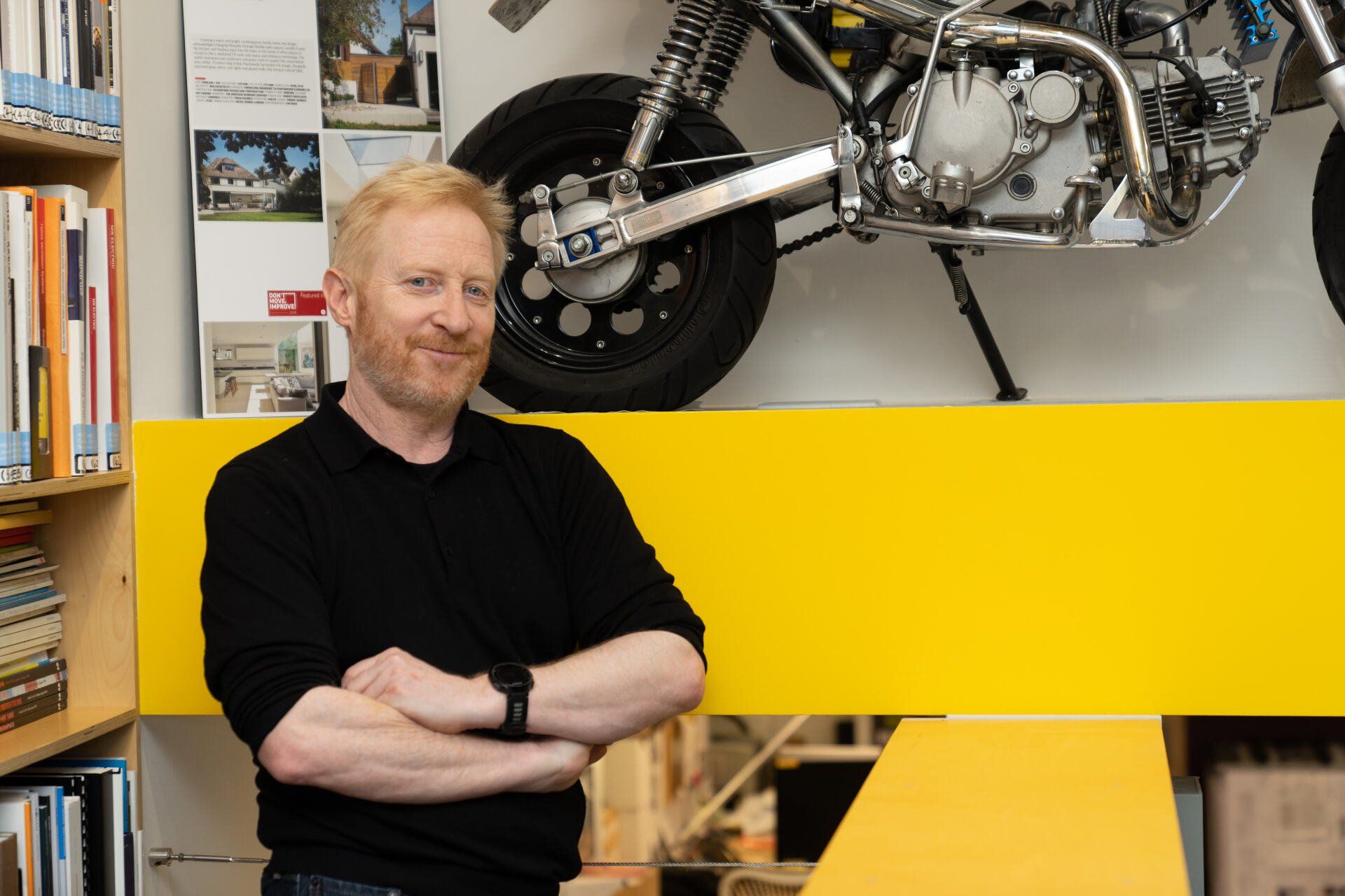 Man standing with arms crossed in a workshop beside a motorcycle over a bright yellow panel