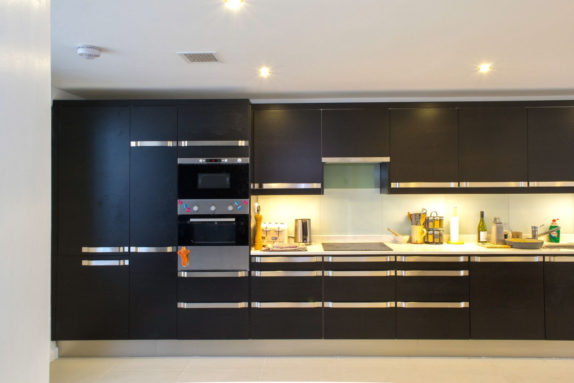 Modern kitchen with black cabinets, built-in oven, and illuminated countertop with bottles and utensils