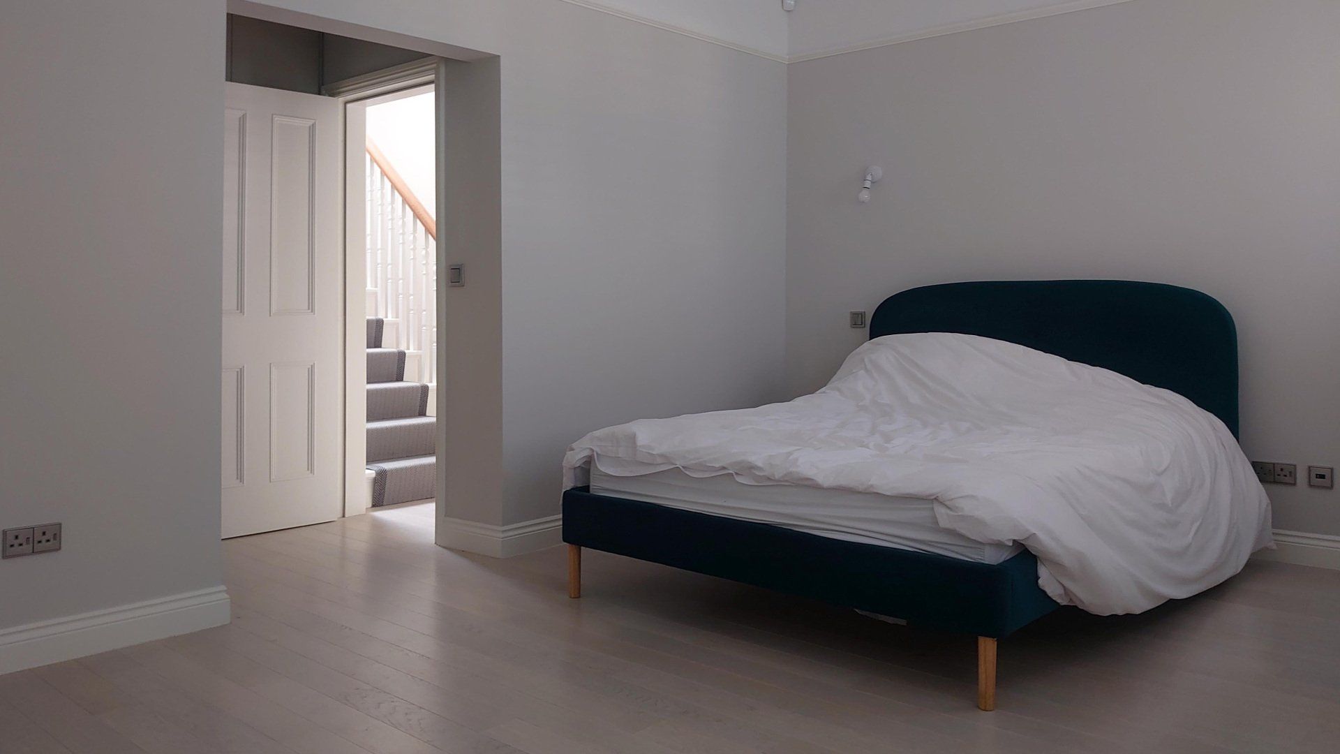 Minimal bedroom with a dark teal bed, white bedding, gray walls, and an open door to a stairway.