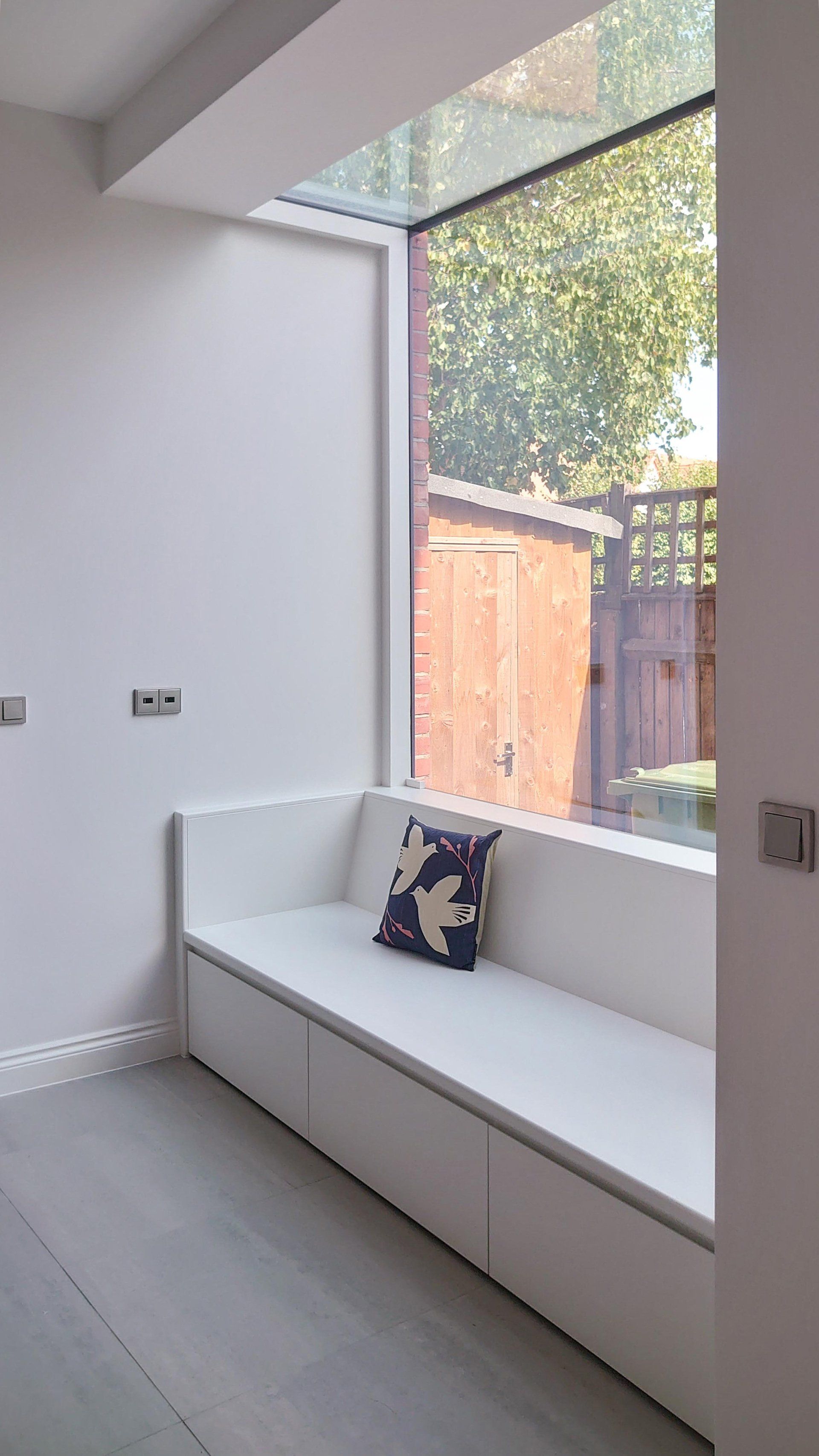 Bright minimalist room with a white bench beneath a large window and a dark patterned cushion.