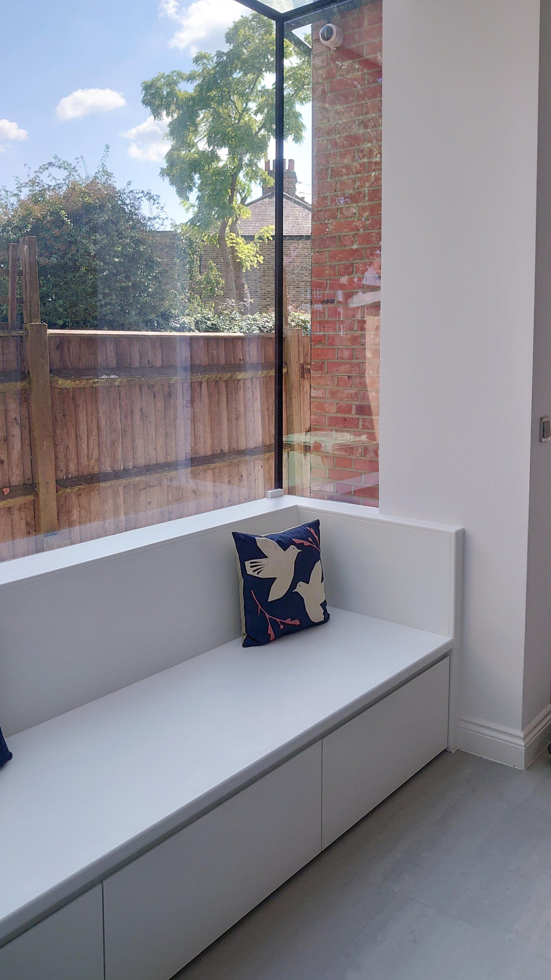 White built-in window seat with a patterned cushion beside a large window overlooking a fenced yard.