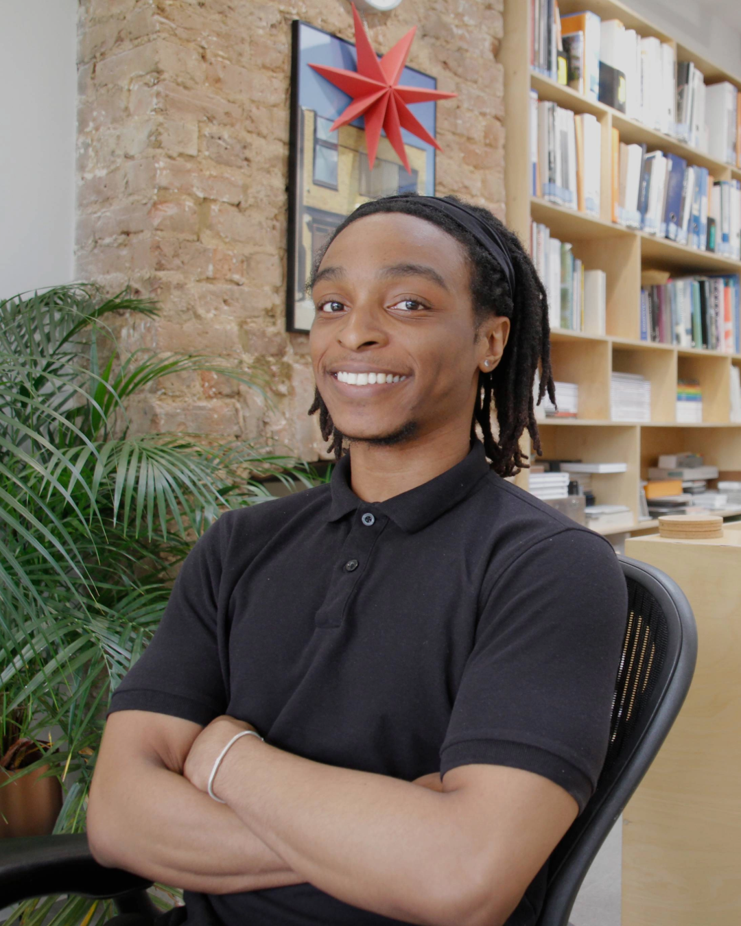 Smiling person with crossed arms seated in a library, with bookshelves and a red star wall decoration behind them
