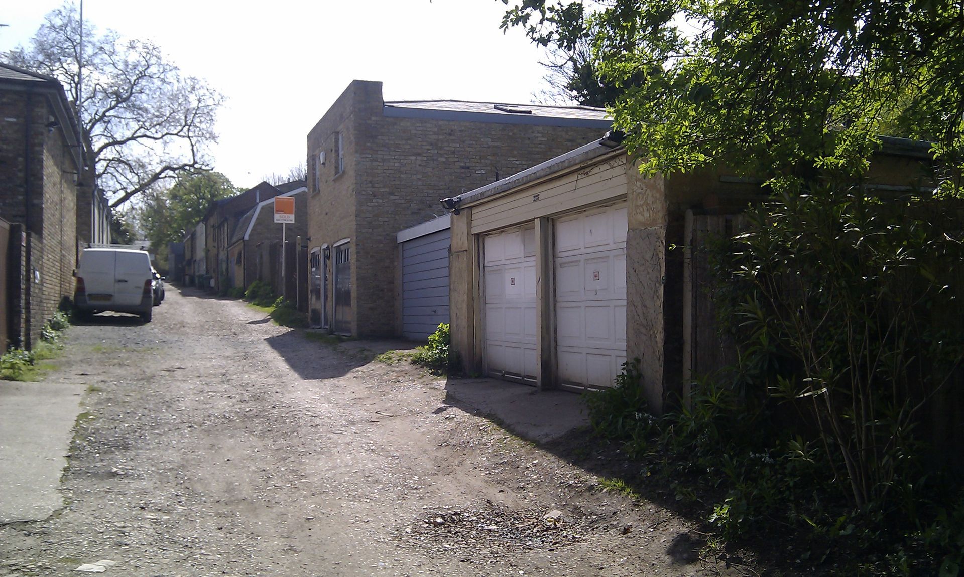 Narrow gravel alley with brick garages, a parked white van, and trees casting shade