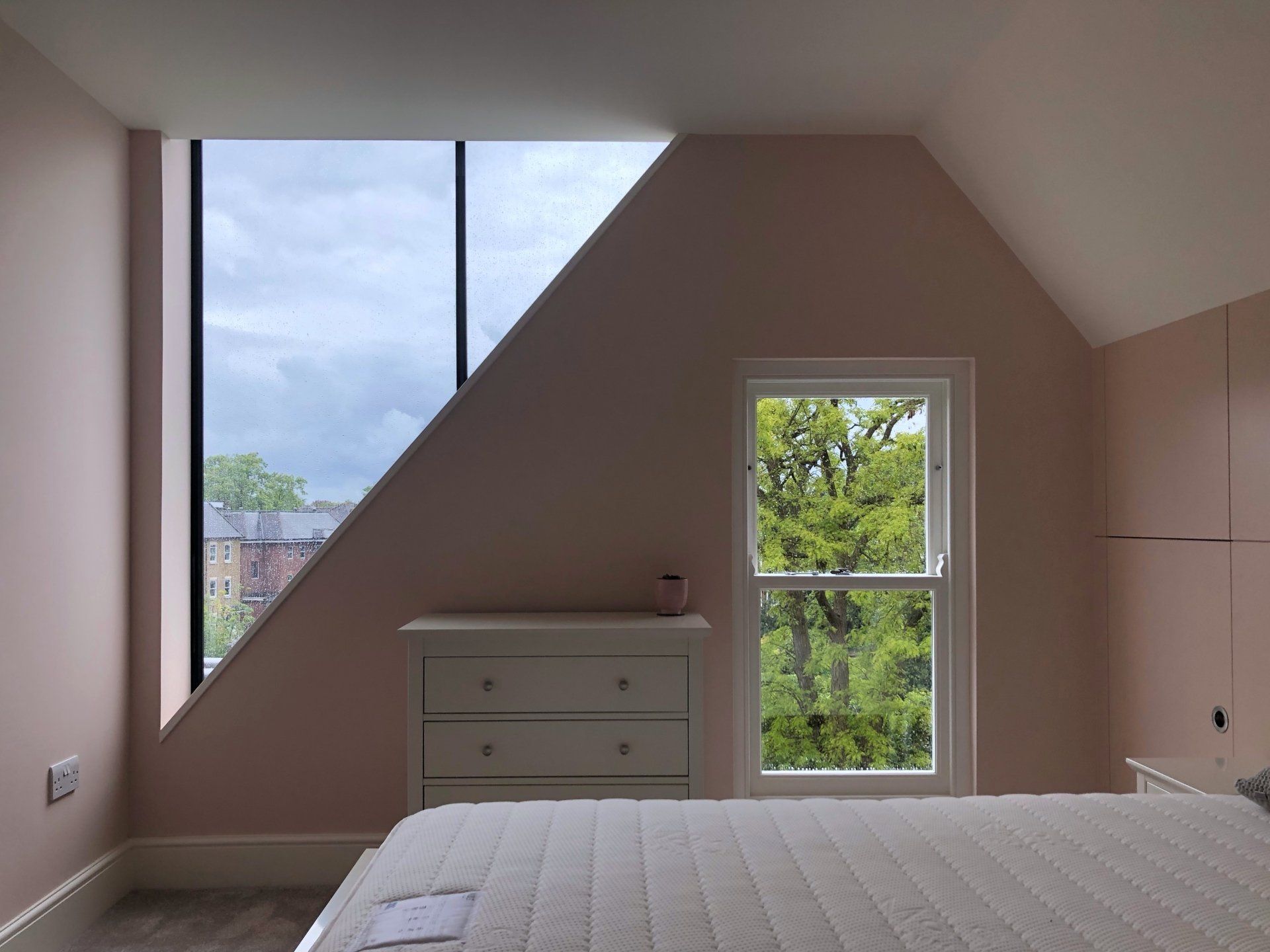 Empty attic bedroom with sloped ceiling, large window, and small dresser beside a glass door.