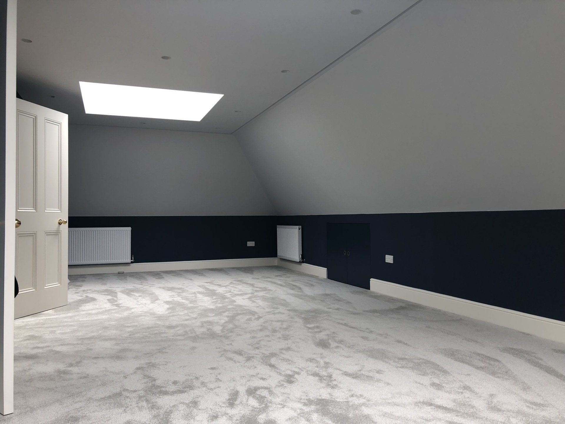 Empty attic bedroom with gray walls, dark blue wainscoting, and white carpet under a skylight.