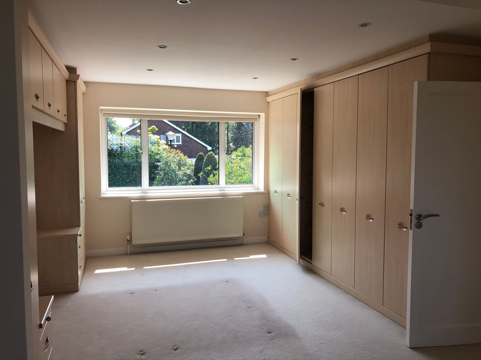 Empty beige bedroom with built-in wardrobes, open door, and window overlooking greenery