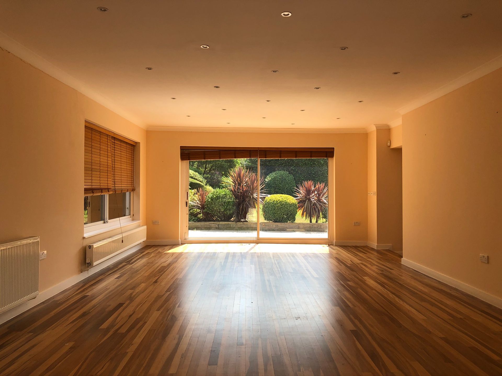 Empty living room with wooden floors and large sliding doors opening to a sunny garden.