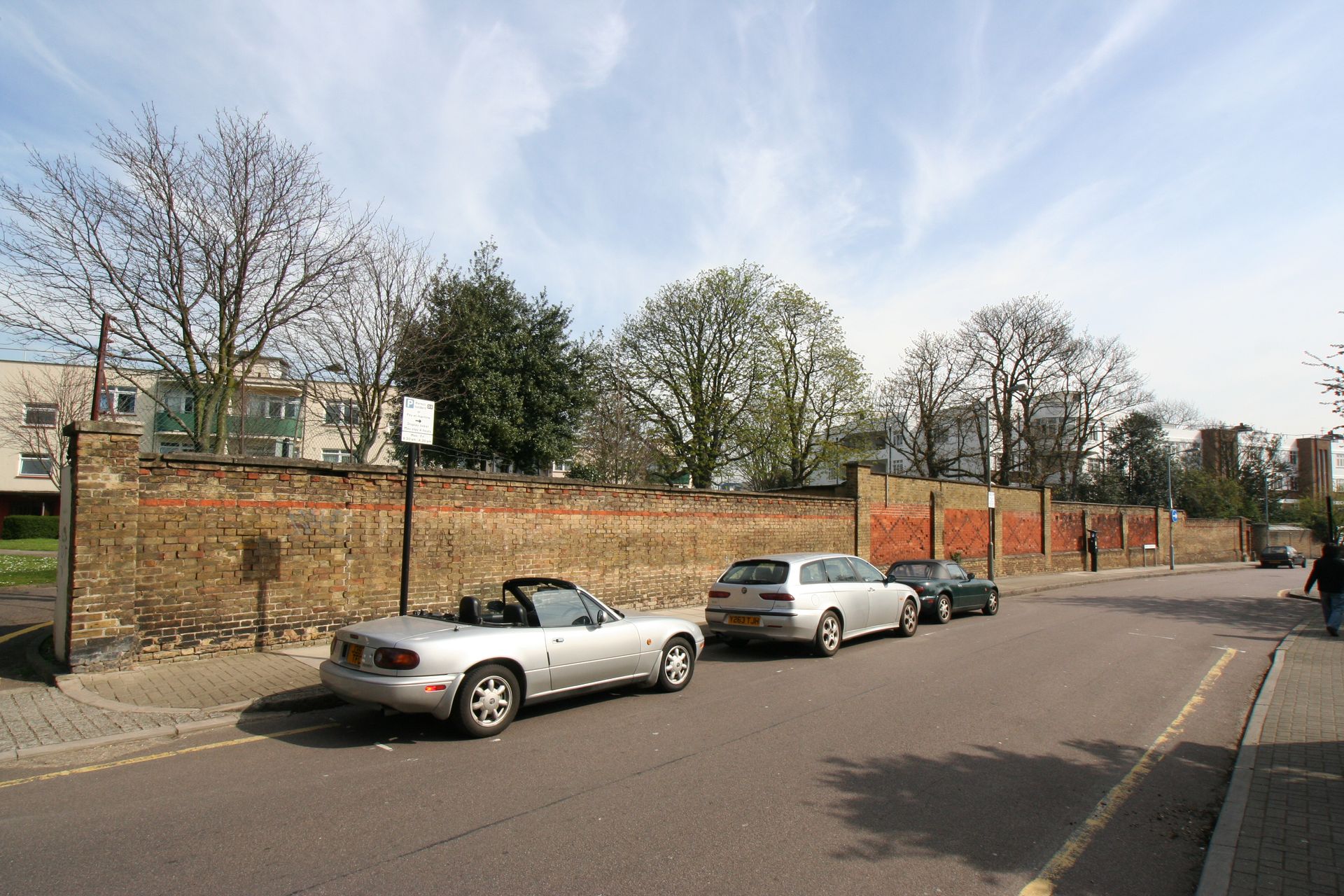 Parked cars along a brick wall on a quiet street under a partly cloudy sky.