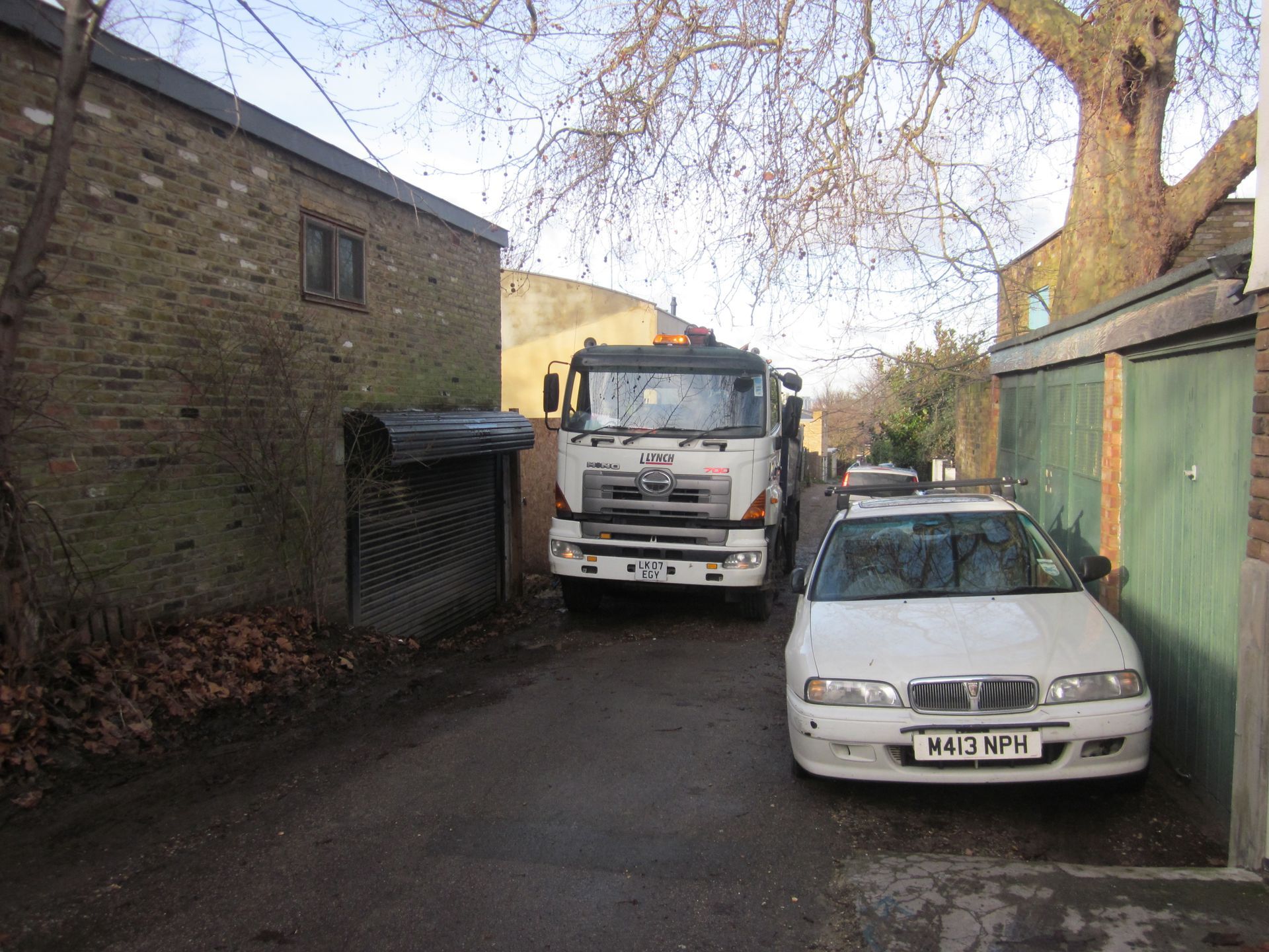 Narrow alley with a white truck blocked by a parked white car between brick walls and green garage doors.