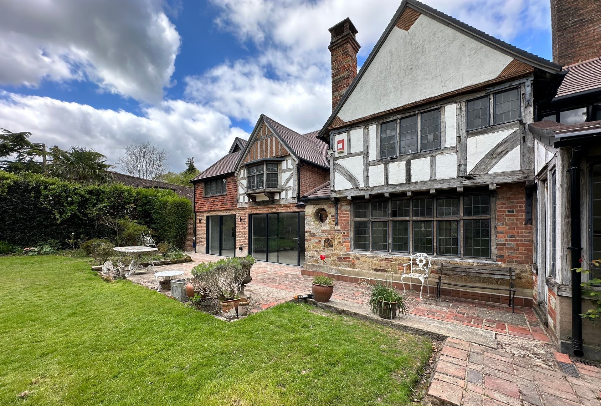 Stone-and-timber cottage with a brick patio, small garden, and green lawn under a cloudy sky.