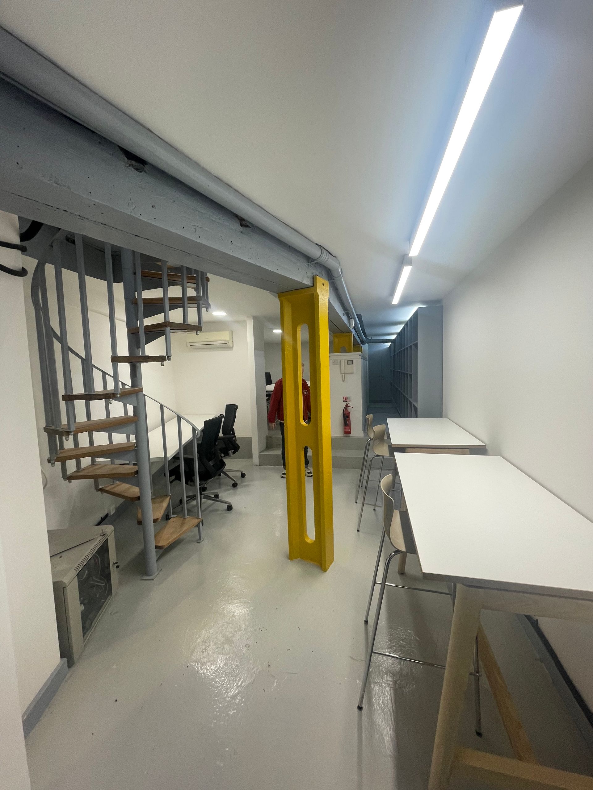 Narrow industrial hallway with stairs, a yellow gate, chairs, and bright overhead lighting.