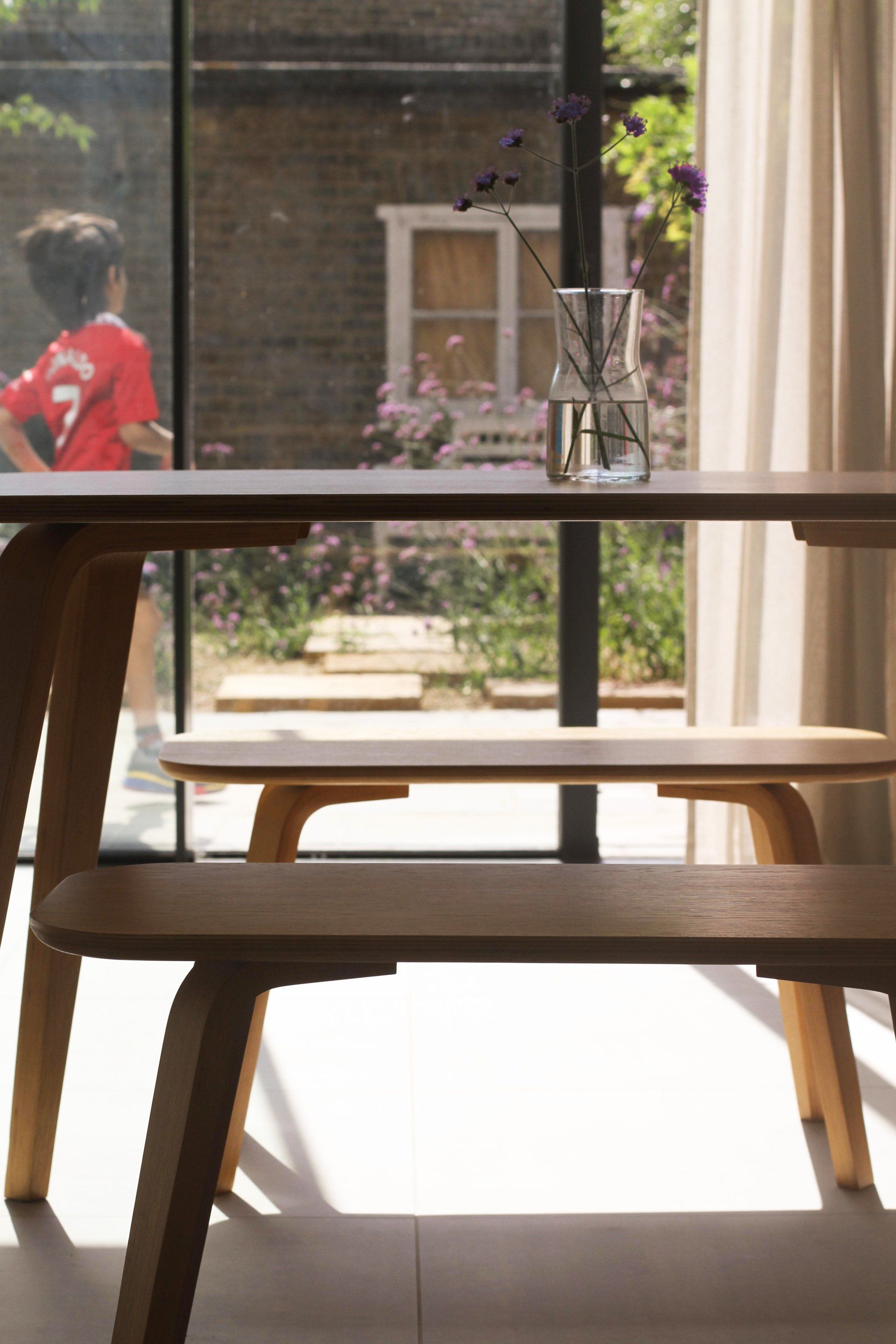 Wooden chairs by a sunlit window overlooking a patio and garden, with a person passing outside