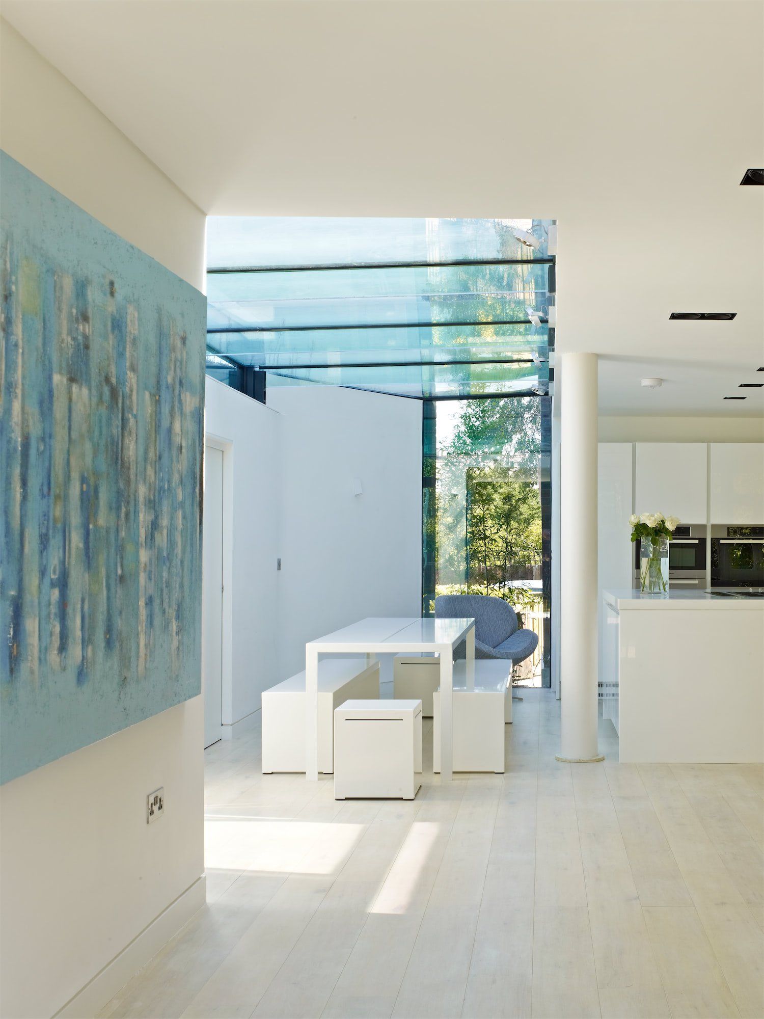 Modern white dining area with glass wall, white table and stools, and abstract blue artwork on the left wall
