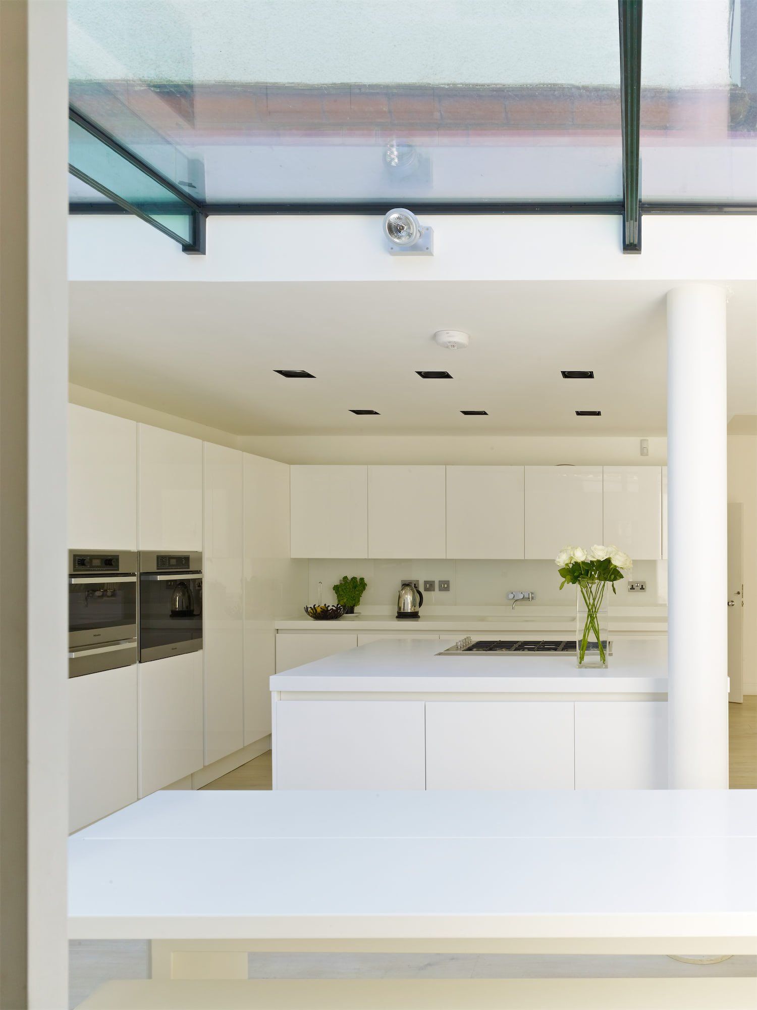 Modern white kitchen with island, built-in ovens, and skylight above slim black-framed windows