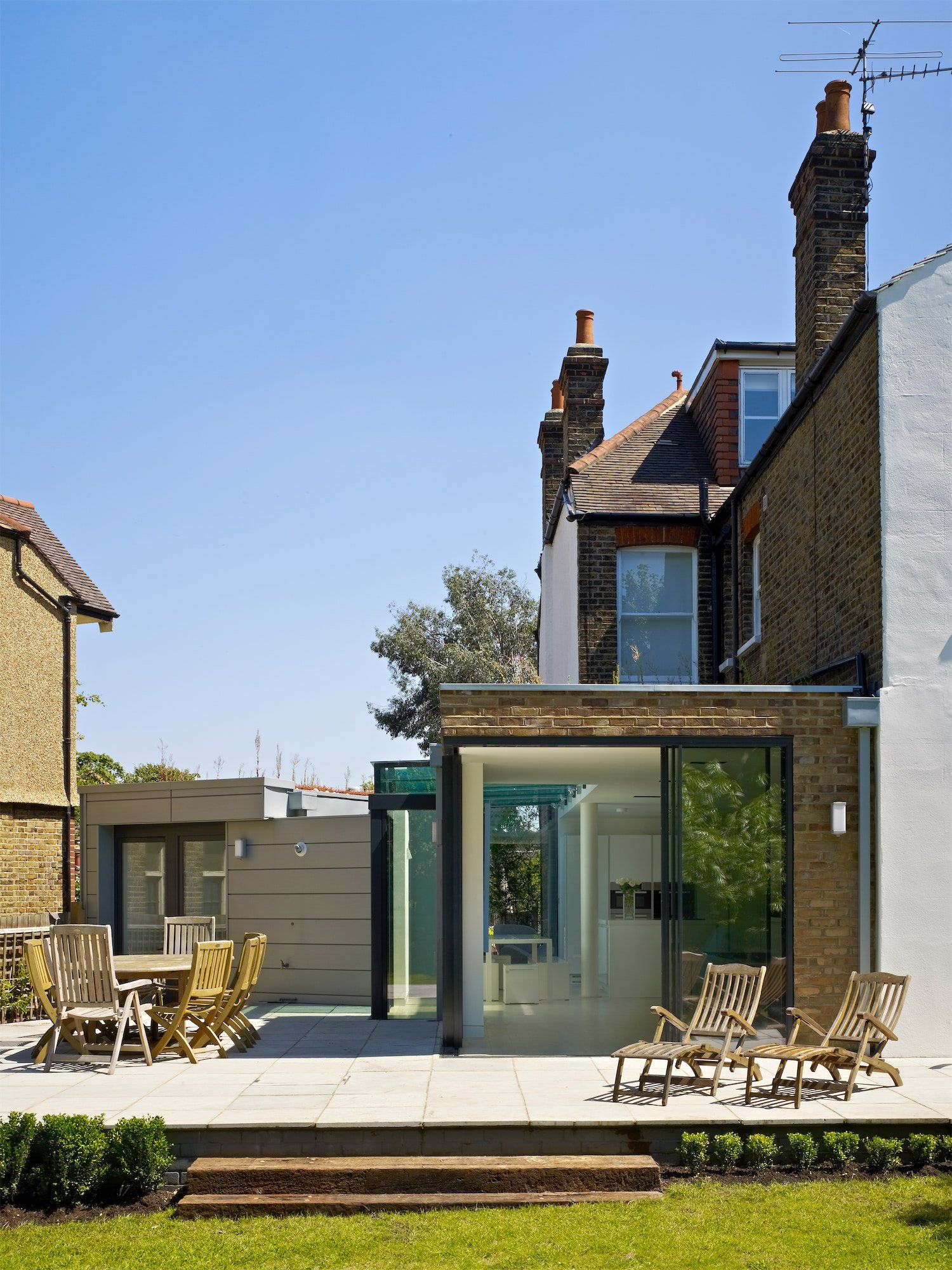 Modern brick house with glass extension, patio chairs, and a garden under a clear blue sky