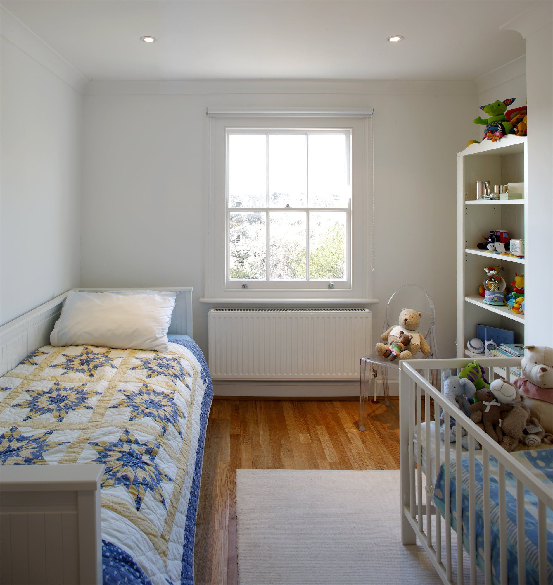 Small, bright children's bedroom with two beds, window, and white shelving unit filled with toys.