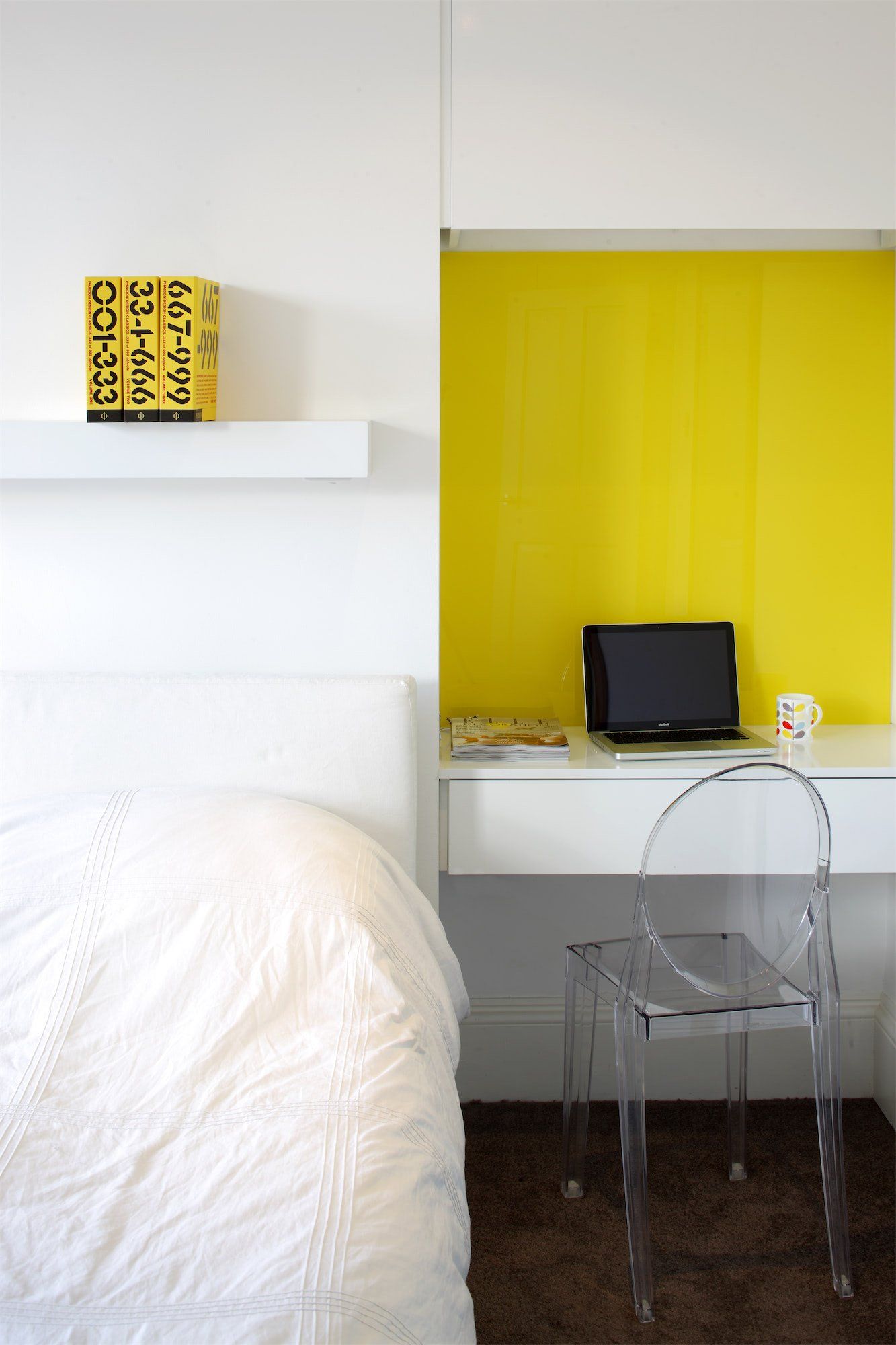 Bright yellow desk area with laptop, clear chair, and white bed.