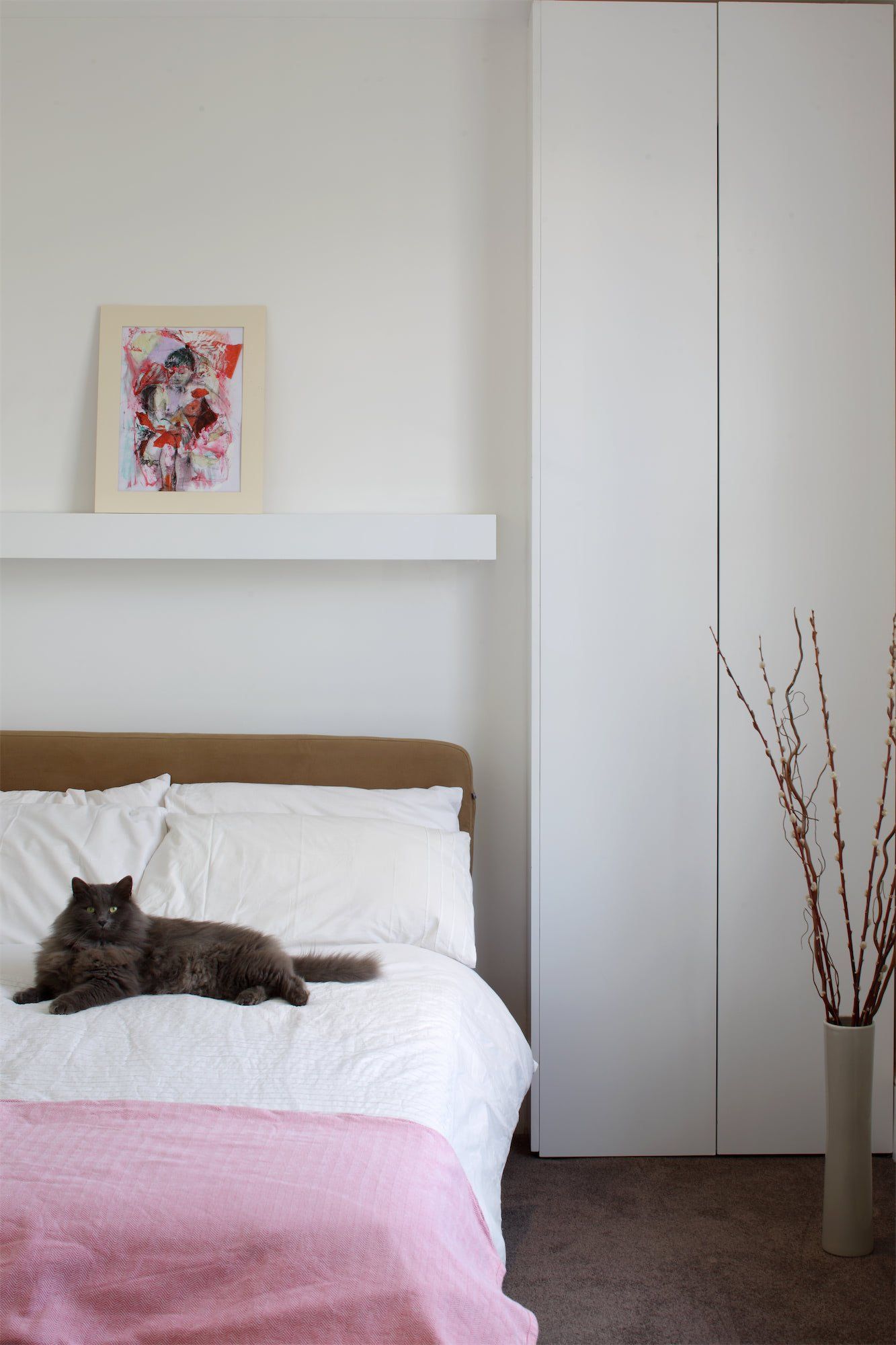 Gray cat on a bed with white bedding, pink blanket, and art, next to white closet.