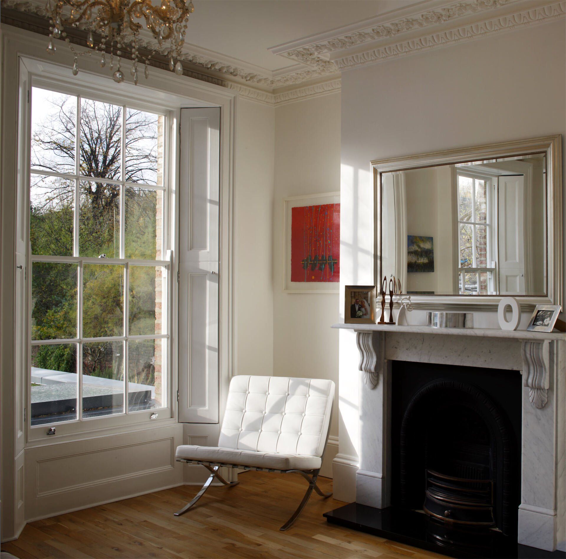 Interior of a bright room with a large window, fireplace, chair, and mirror. White walls and wood flooring.