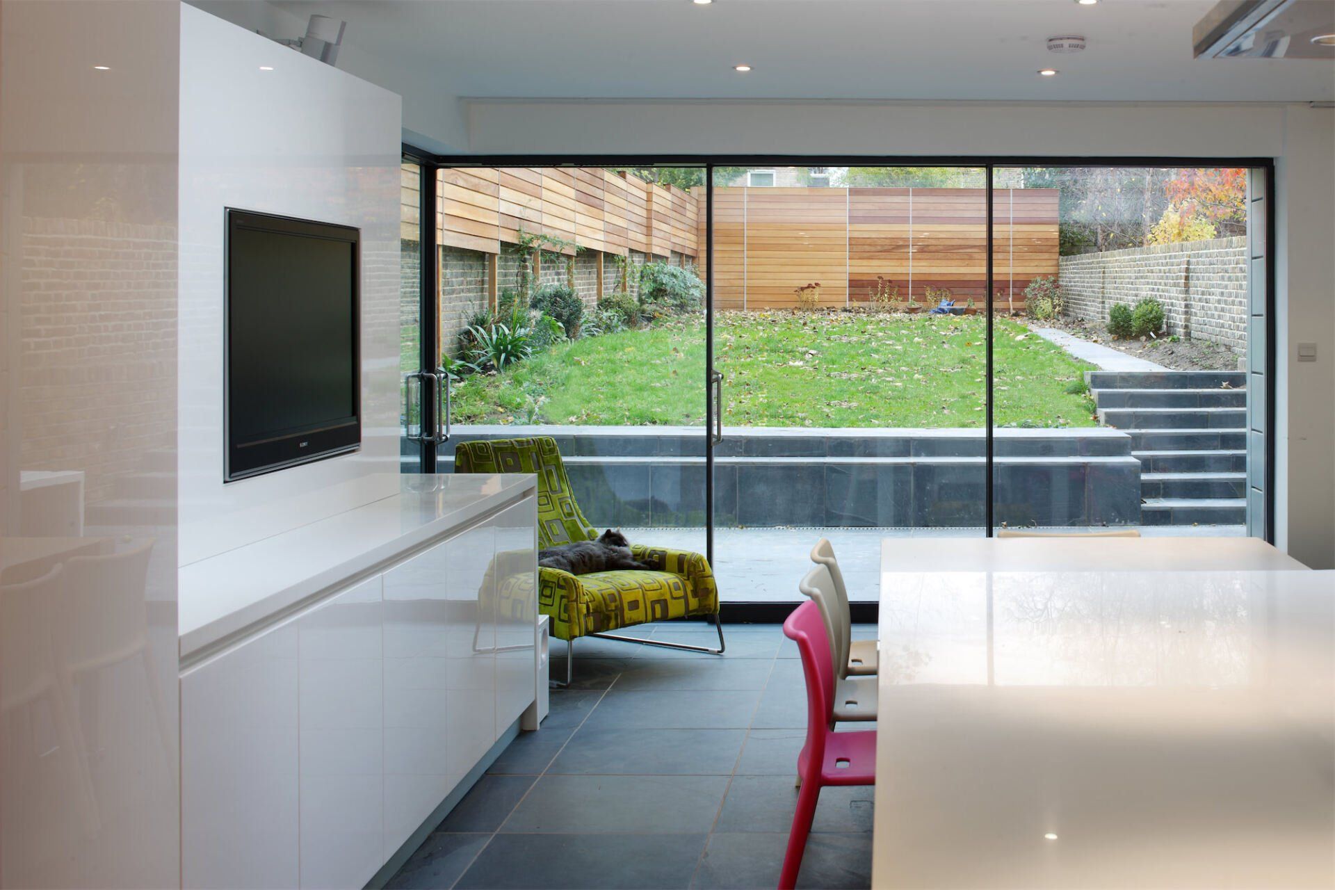 Modern kitchen with white cabinets, dark tile floor, large window overlooking a garden with lawn and wood fence.