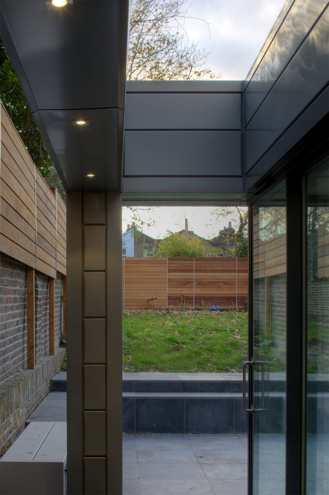 Exterior view: modern gray and brick building opening onto a lawn. Sliding glass door, wooden fence, cloudy sky.