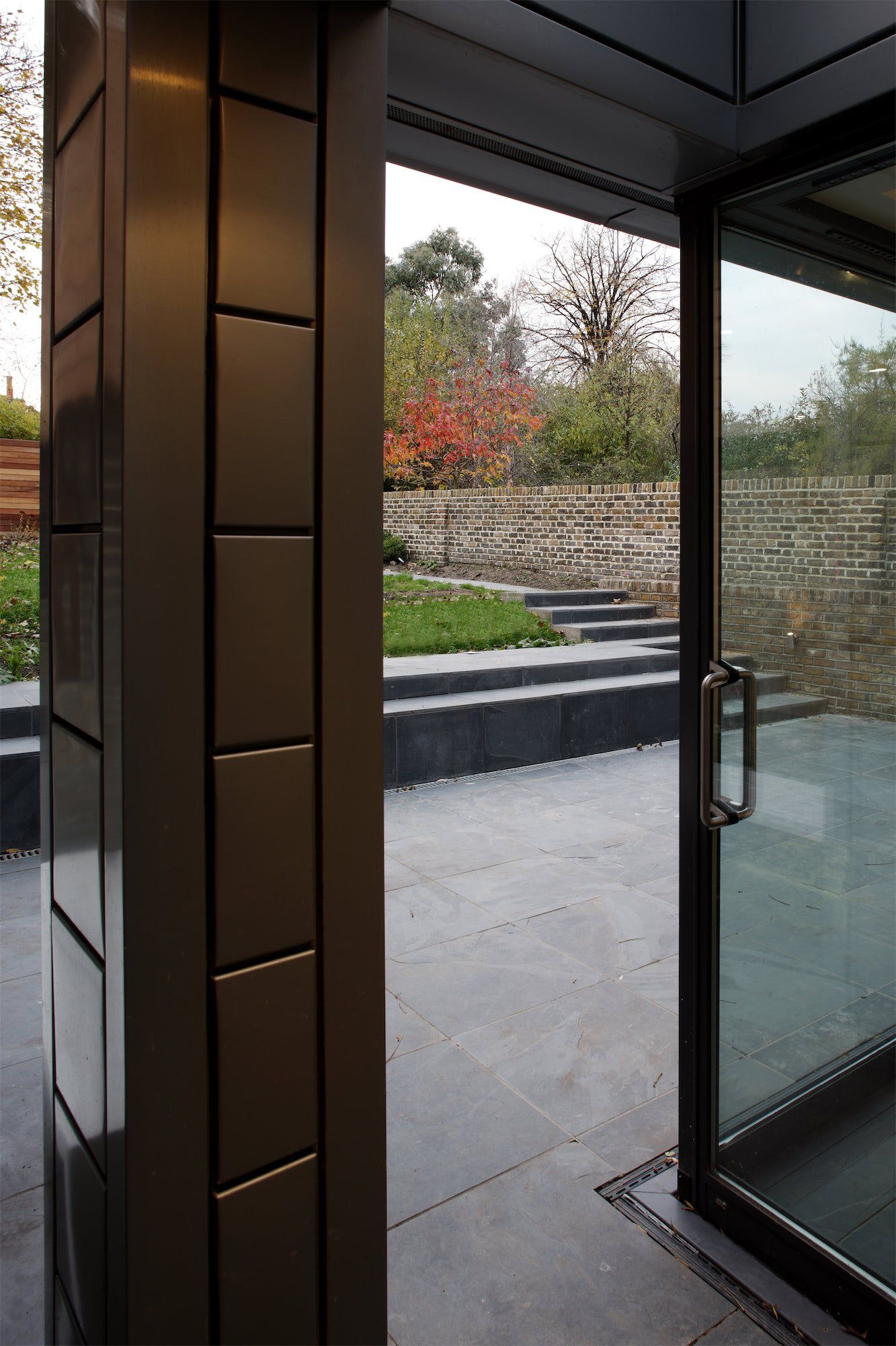 Modern doorway with tiled pillar, open to patio and garden with stone steps and trees.