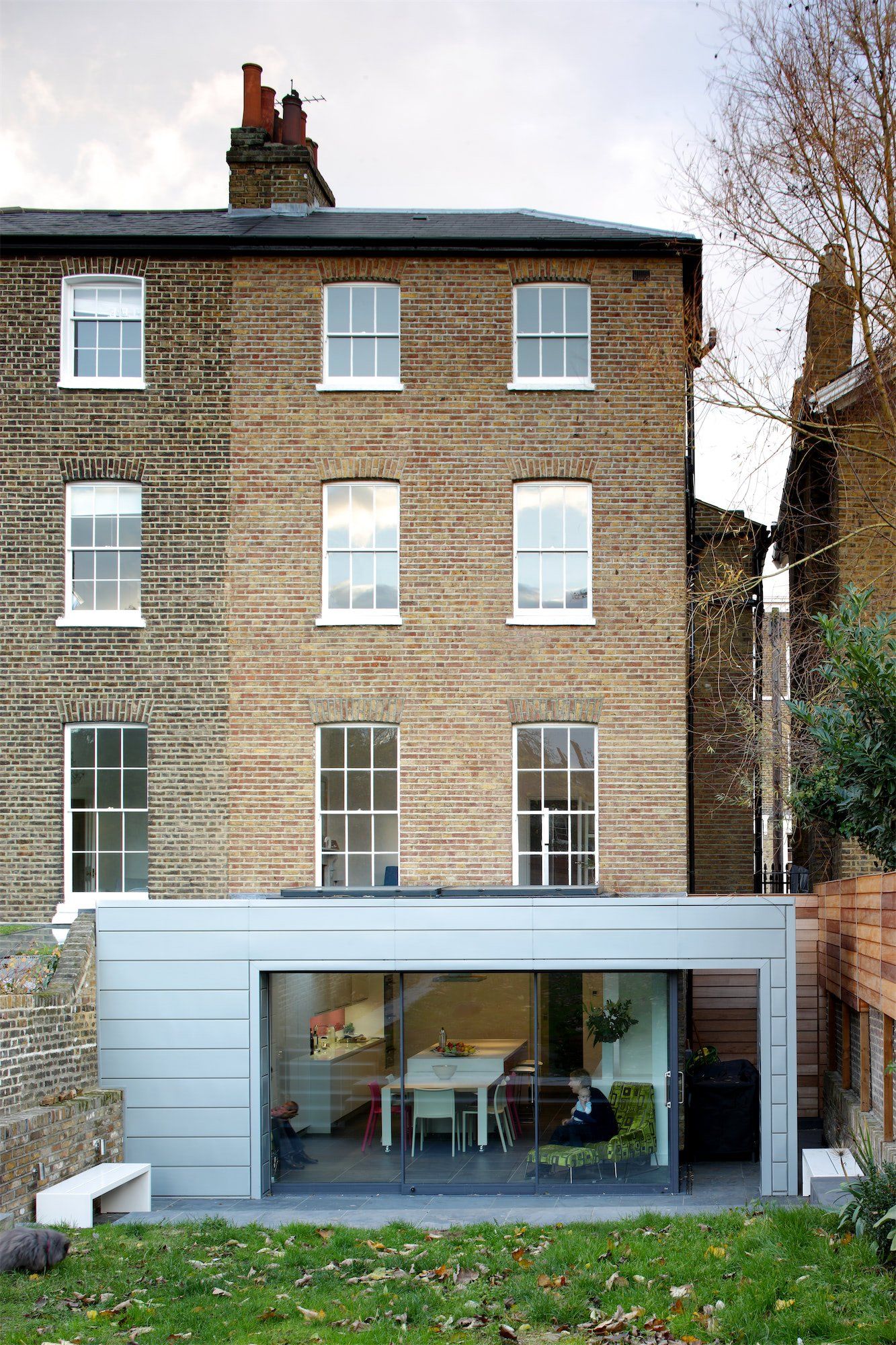 Gray modern addition with glass doors to a brick house, overlooking a green yard and garden.