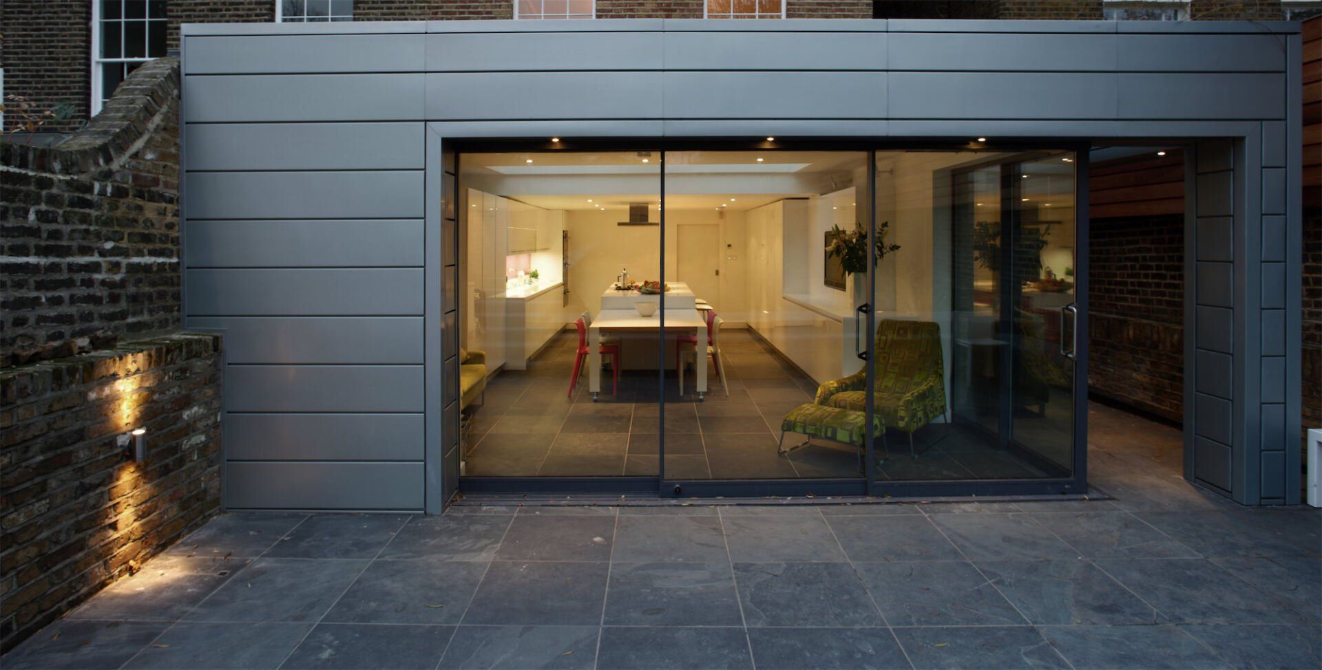 Modern, gray-walled building with glass sliding doors, revealing a well-lit dining area. Dark tiled patio.