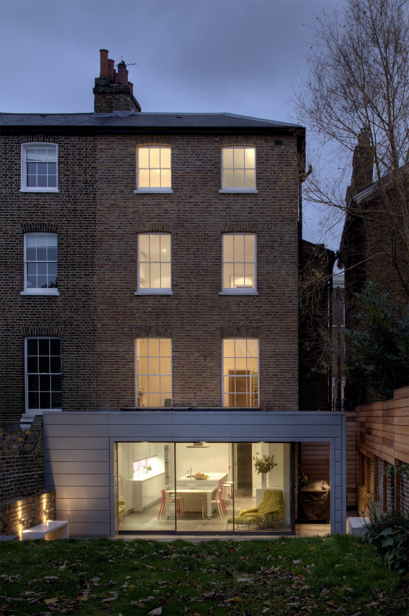 Rear view of a brick townhouse with a modern, glass-walled ground floor extension; illuminated interior.