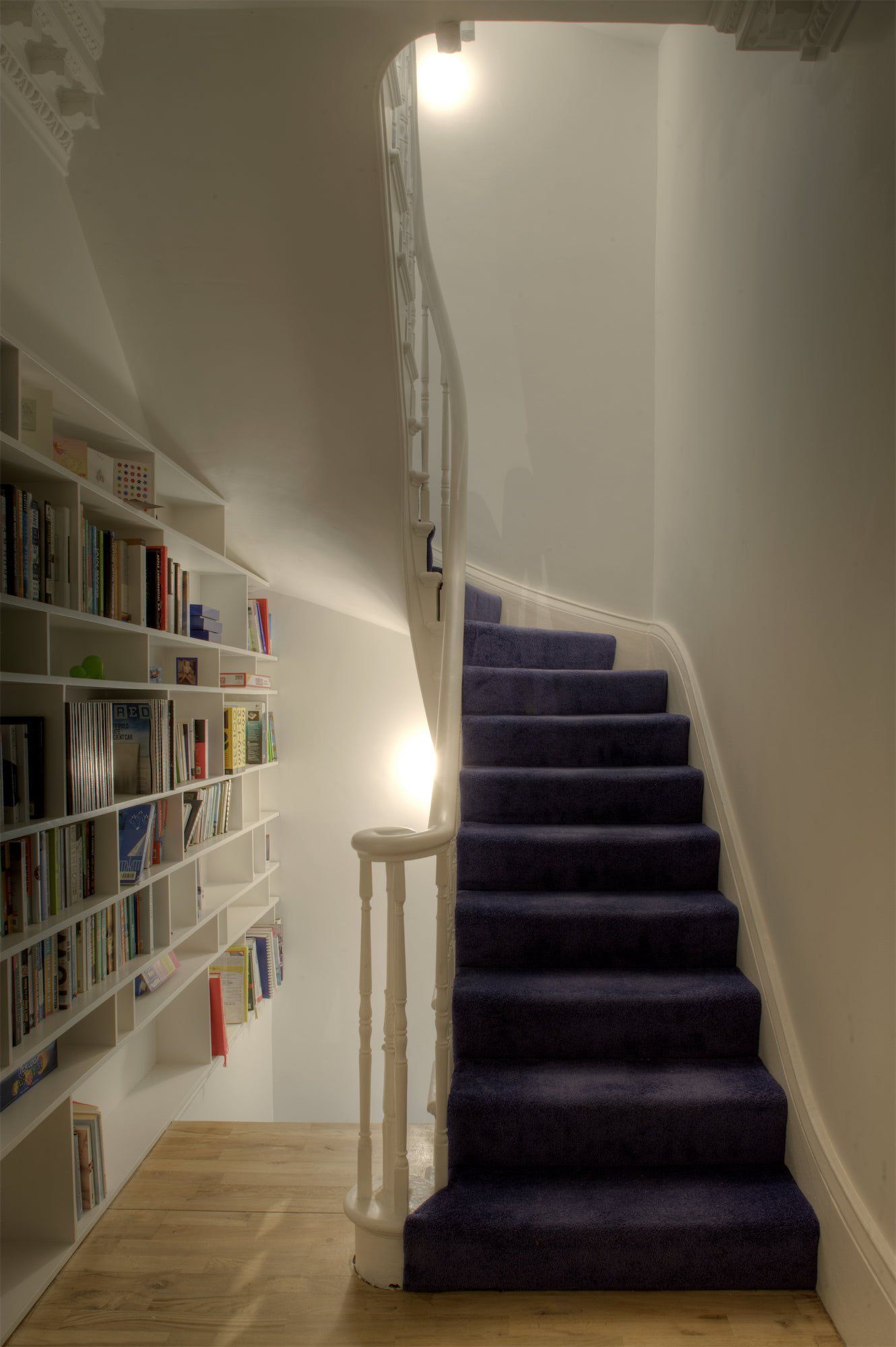 Curving staircase with blue carpet and built-in bookshelf on the wall, lit by soft lighting.