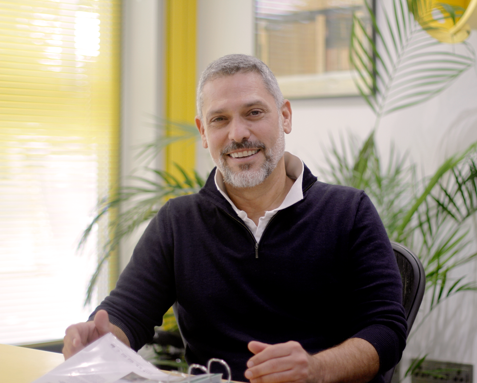 Smiling man seated indoors at a table with plants and yellow curtains in the background